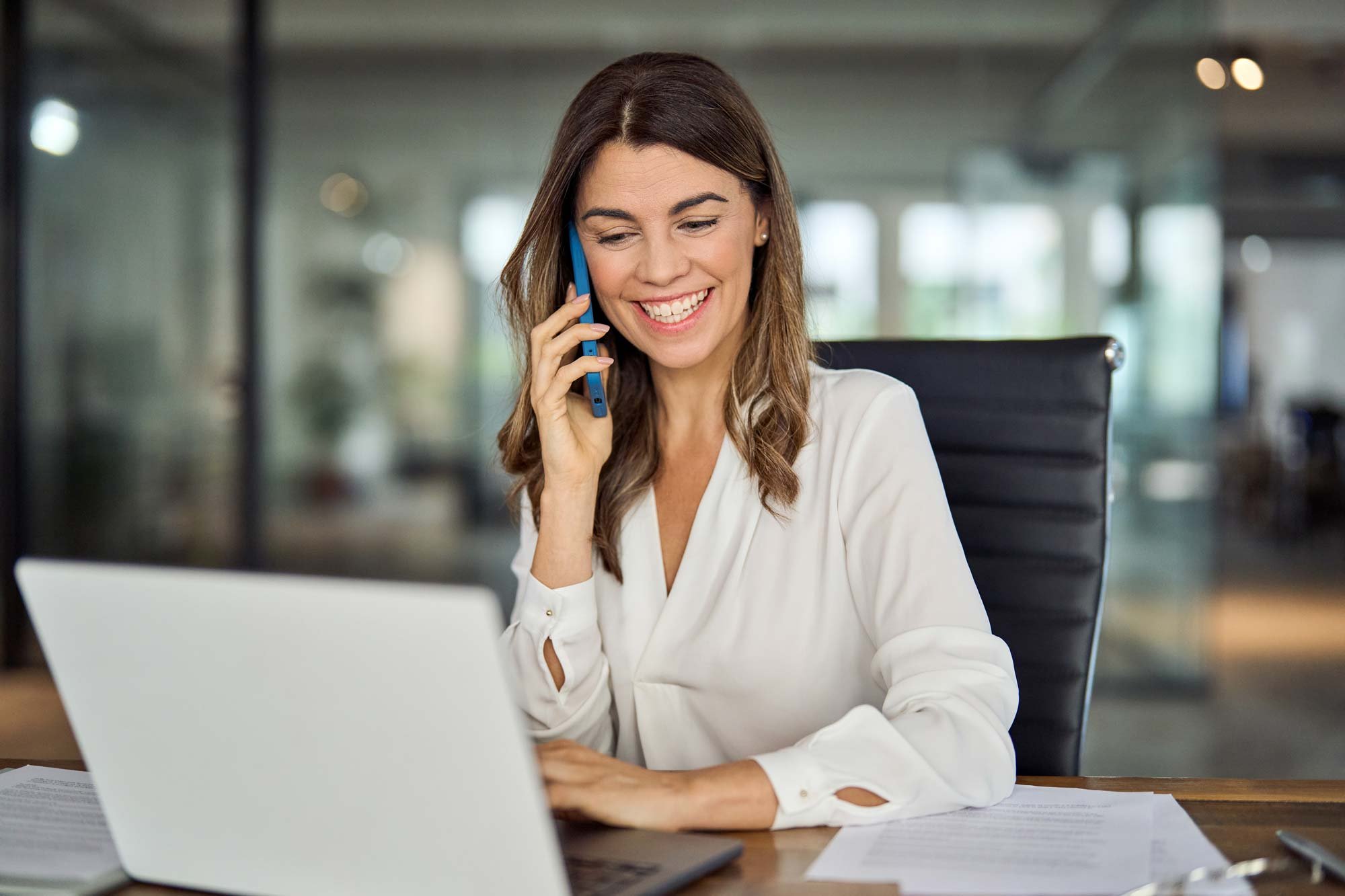 A woman with brown hair smiling and talking on a blue cell phone in an office. She is sitting at a desk with a laptop and papers, wearing a white blouse.