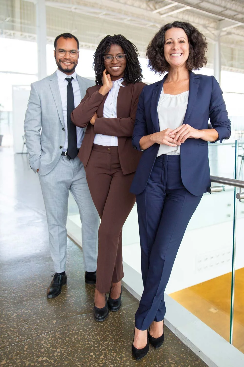 Three professionally dressed businesswomen standing in an office building, smiling at the camera.