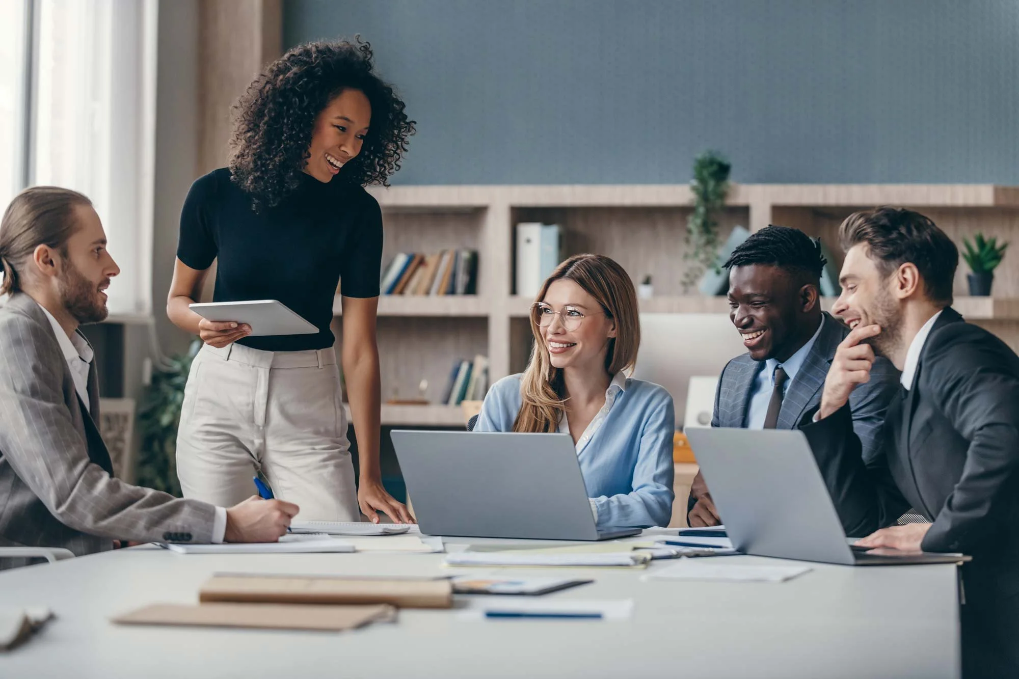 A diverse group of five young professionals is gathered around a table in a modern office, engaged in a discussion. Four are seated with laptops and notebooks, while one woman stands holding a tablet, smiling and talking to the group.