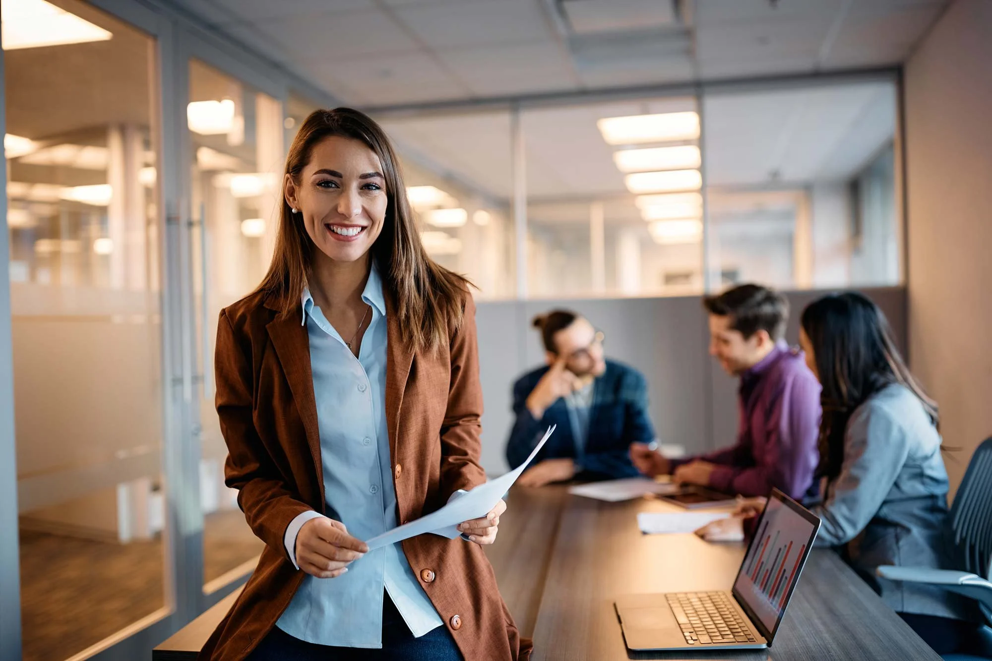 Smiling woman in business attire holding documents in a conference room with three colleagues in the background.