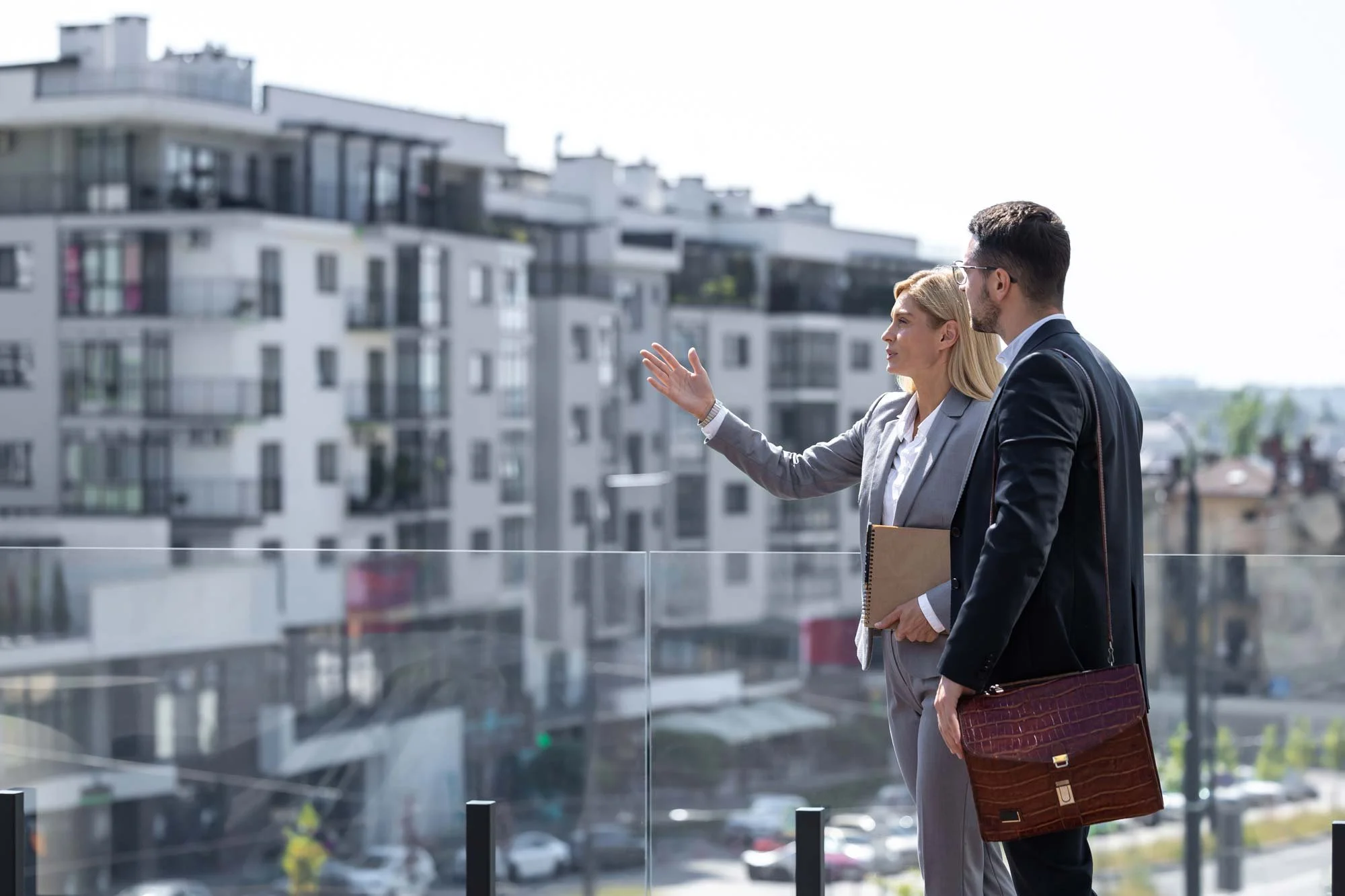 Two business professionals, a woman and a man, discussing on a balcony overlooking apartment buildings in an urban area during daytime.