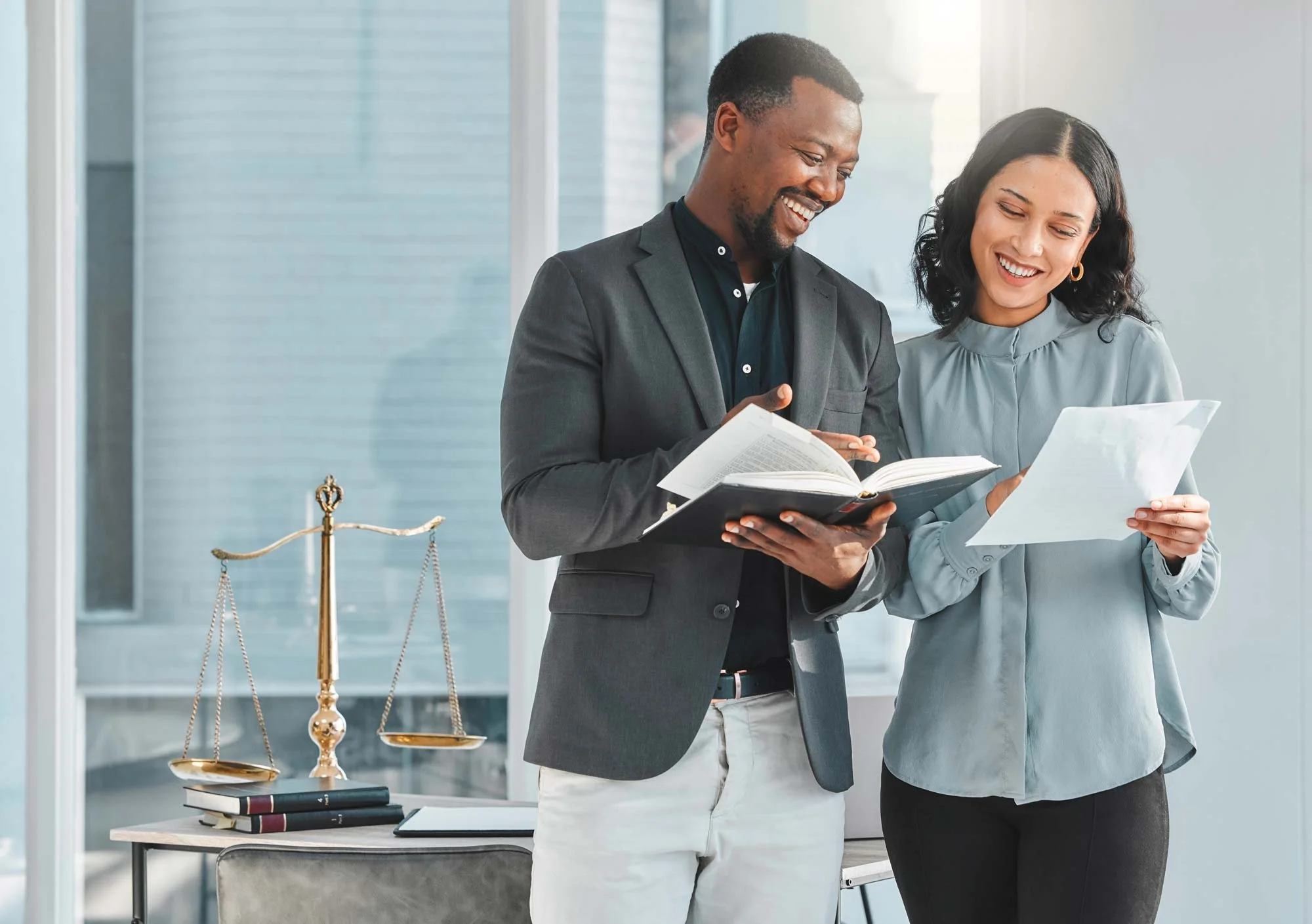 A man and a woman standing together, smiling and looking at documents in a bright office. The man is holding a book, and the woman is holding a sheet of paper. There are legal scales and books on a desk nearby.