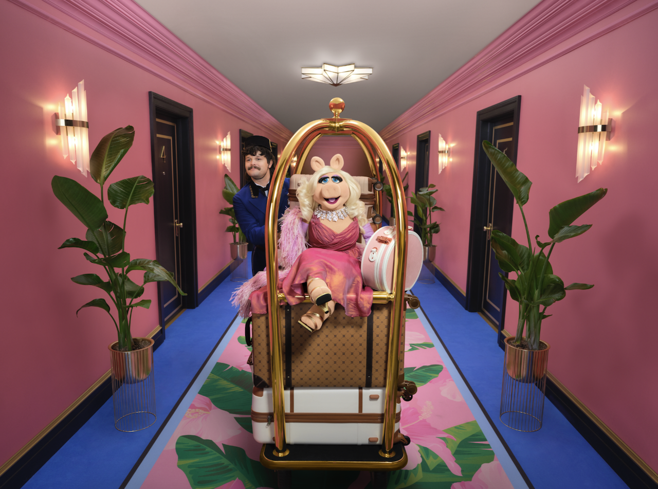 Miss Piggy in a pink hotel hallway, being pushed on a luggage cart by a bell hop.