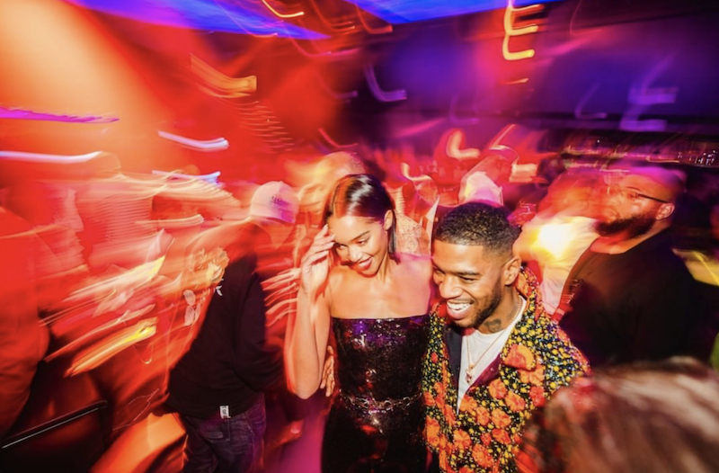 A bcolor photo of two people in a colorfully lit space, laughing and walking toward the camera