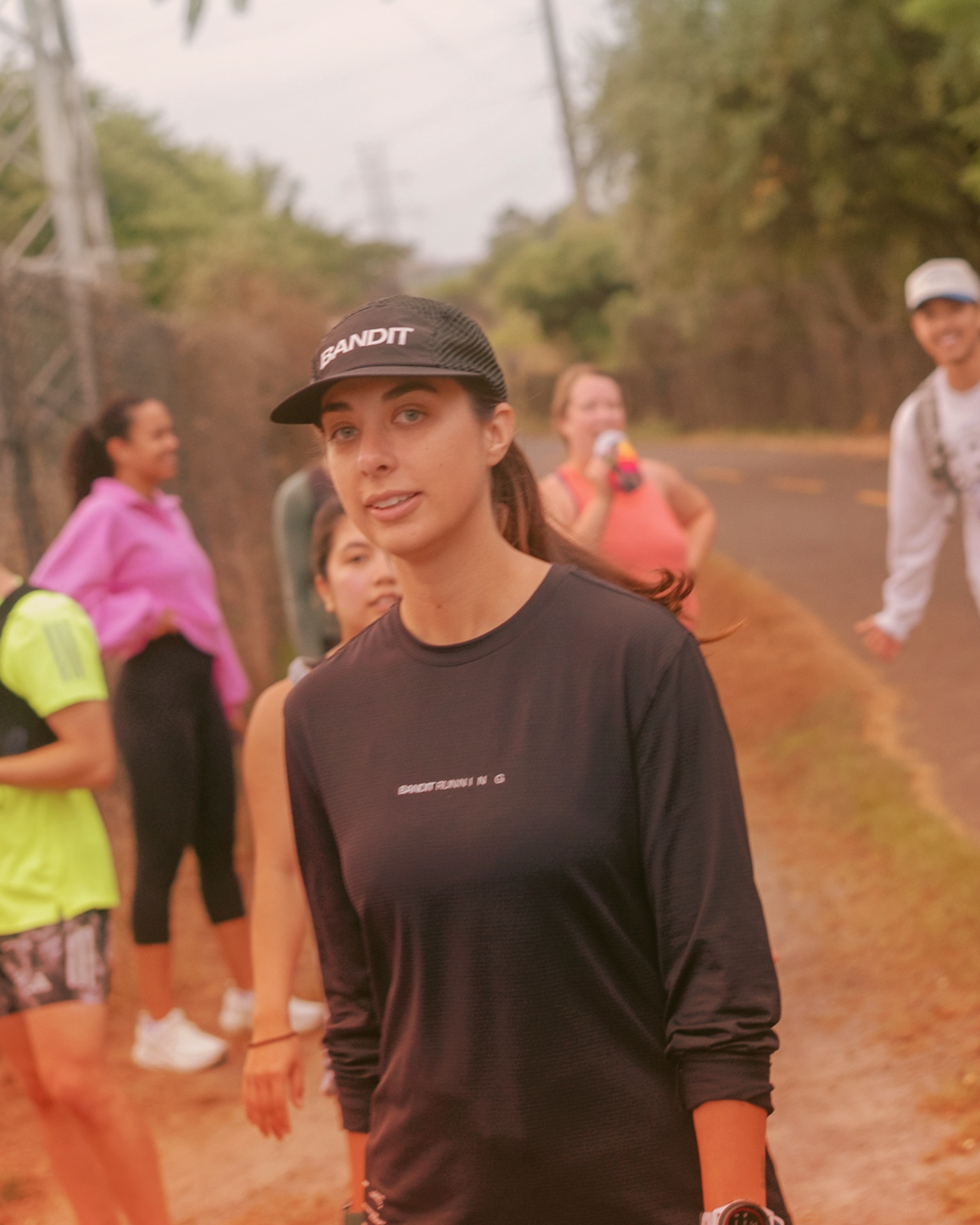 A group of people, including a young woman in the foreground wearing a black long-sleeve shirt and a black cap, standing on a dirt path outdoors during daytime.
