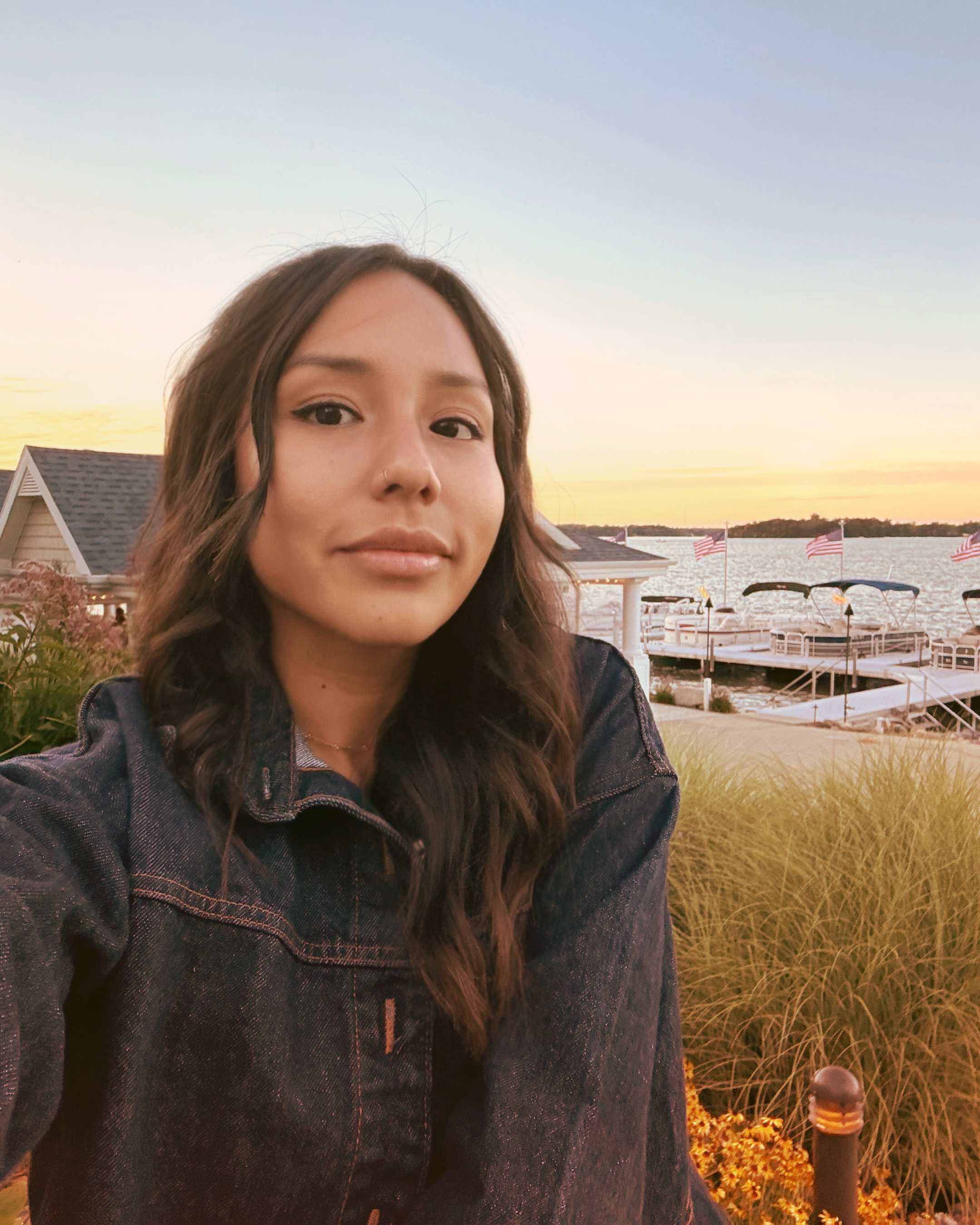 A woman with long wavy brown hair taking a selfie outdoors during sunset by a lakeside with houses, American flags, and boats in the background.