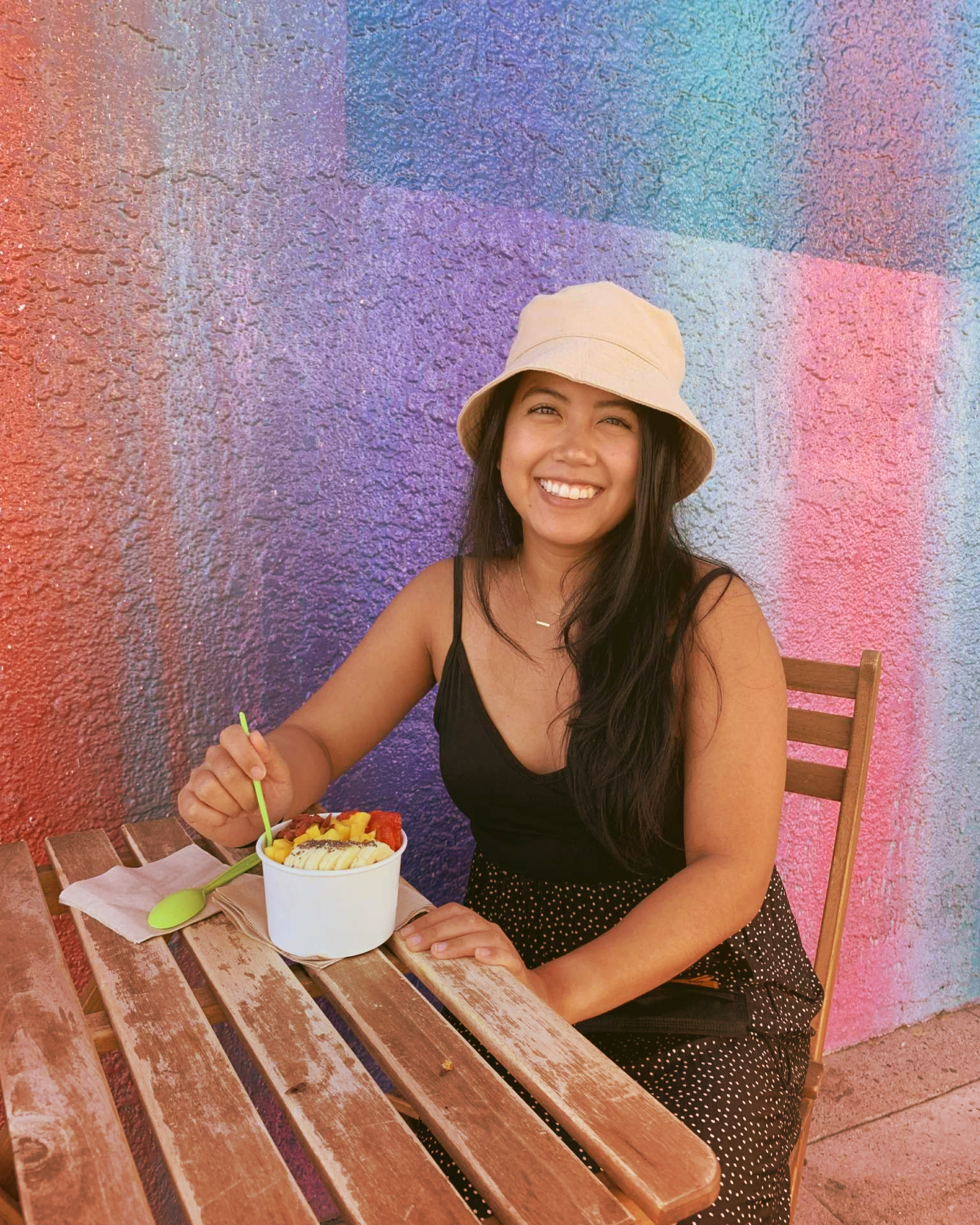 A woman sitting at a wooden table with a bowl of ice cream and fruit, smiling, wearing a beige bucket hat, black sleeveless top, and a black skirt with white dots, against a colorful graffiti wall.