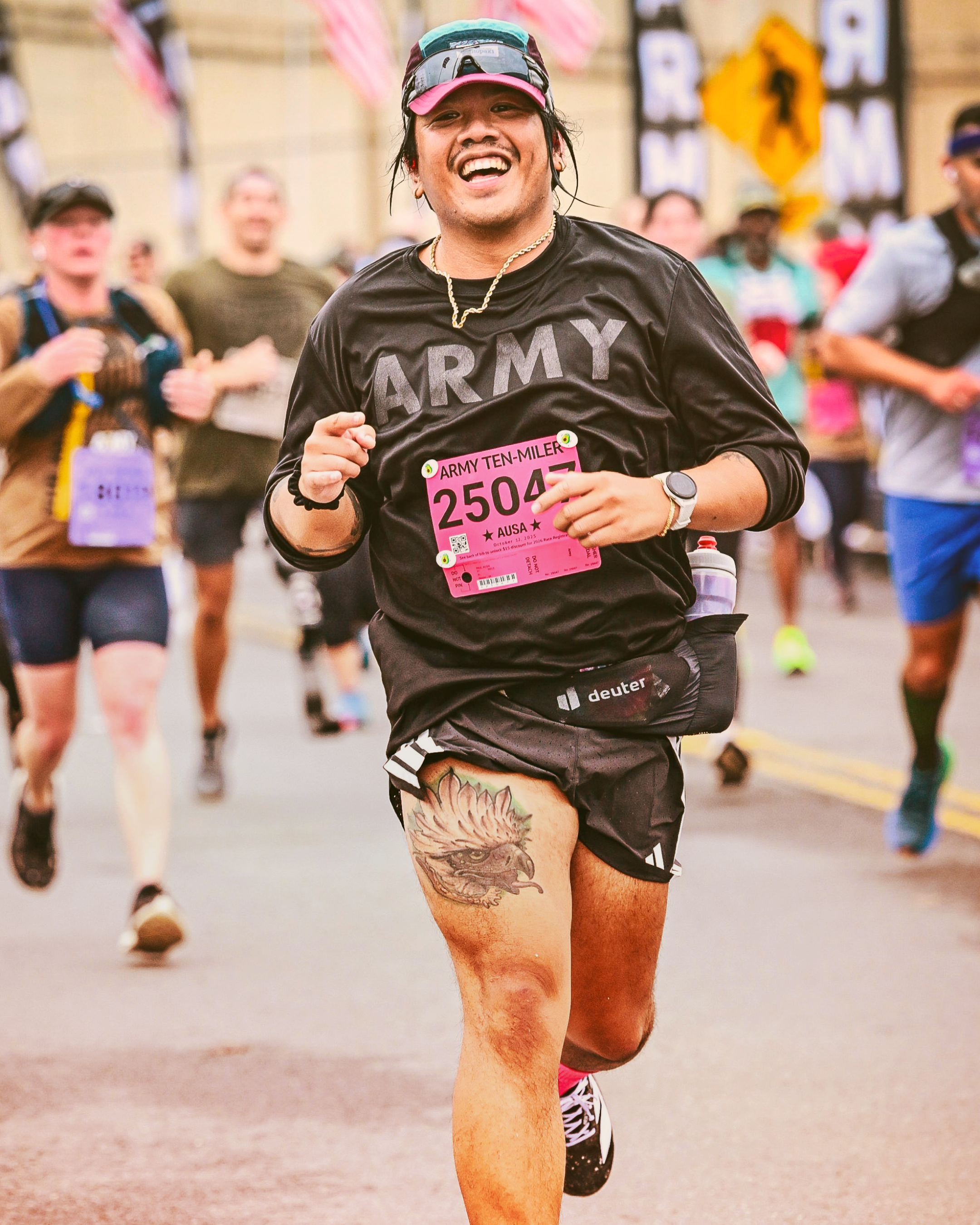 Young man standing indoors, making a peace sign with his right hand. He is smiling and wearing a gray shirt with a design featuring a deer skull and text, and a tan beanie. The background shows a window with blinds and part of a dark room.