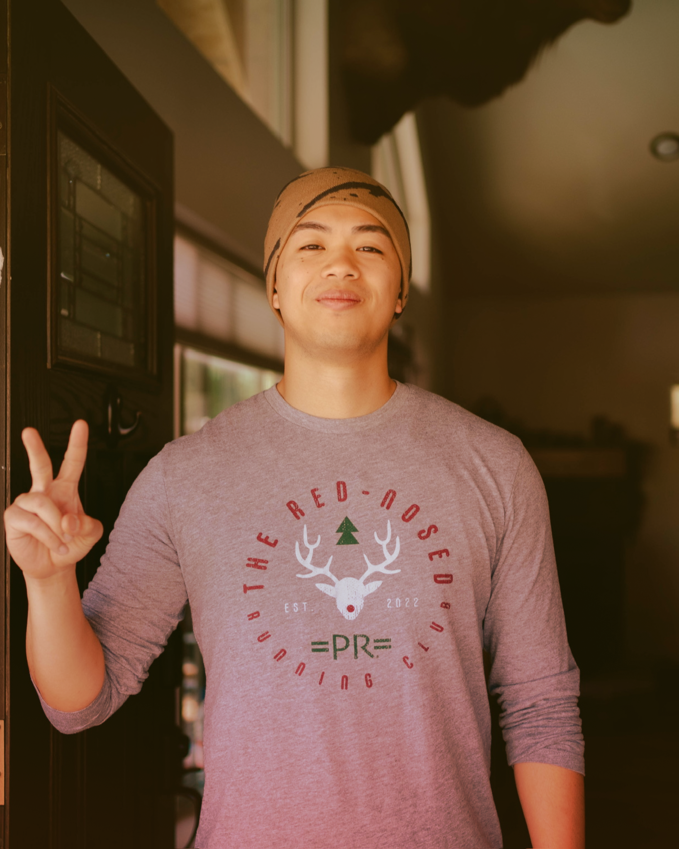 Young man standing indoors, making a peace sign with his right hand. He is smiling and wearing a gray shirt with a design featuring a deer skull and text, and a tan beanie. The background shows a window with blinds and part of a dark room.