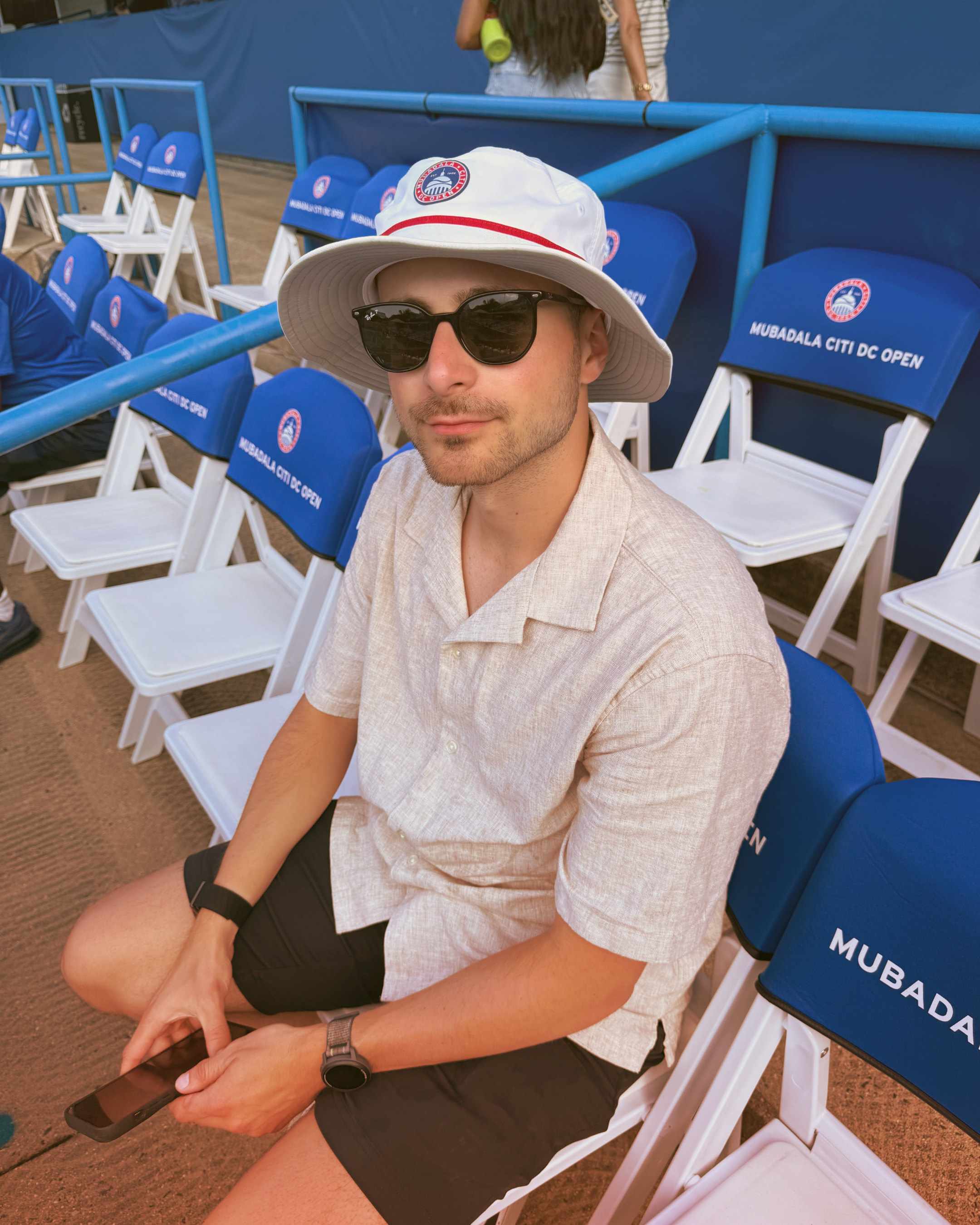 A man sitting on white chairs with blue covers at the MUBADALA CITY DC OPEN tennis event, wearing a white sun hat, black sunglasses, a light-colored short-sleeve shirt, black shorts, and holding a smartphone.