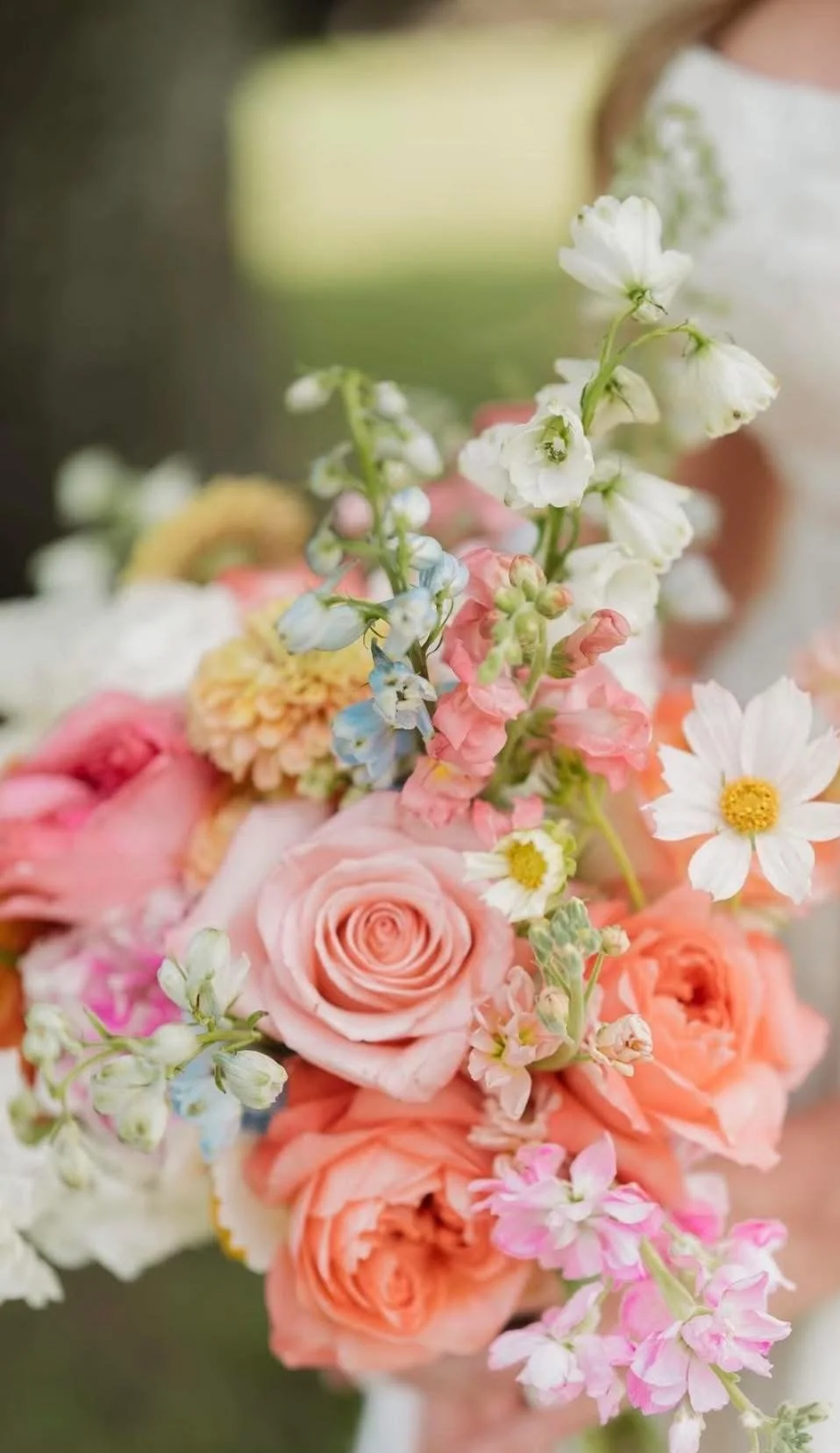 Close-up of a colorful flower bouquet including pink roses, white daisies, blue and pink delphiniums, and other mixed flowers.