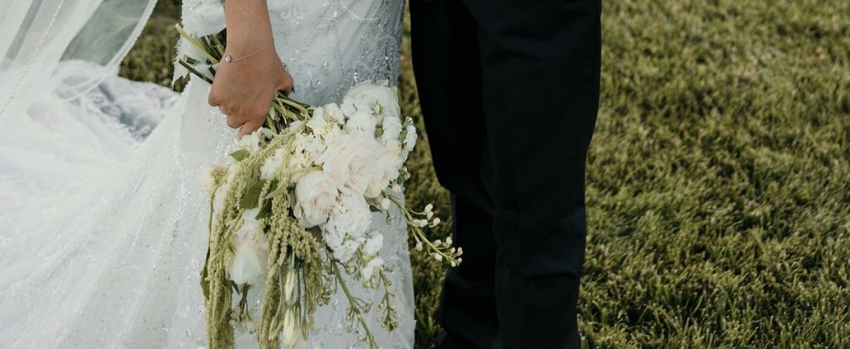 A bride in a wedding dress holding a bouquet of white flowers and greenery, standing next to a groom dressed in black pants, on a grassy field.