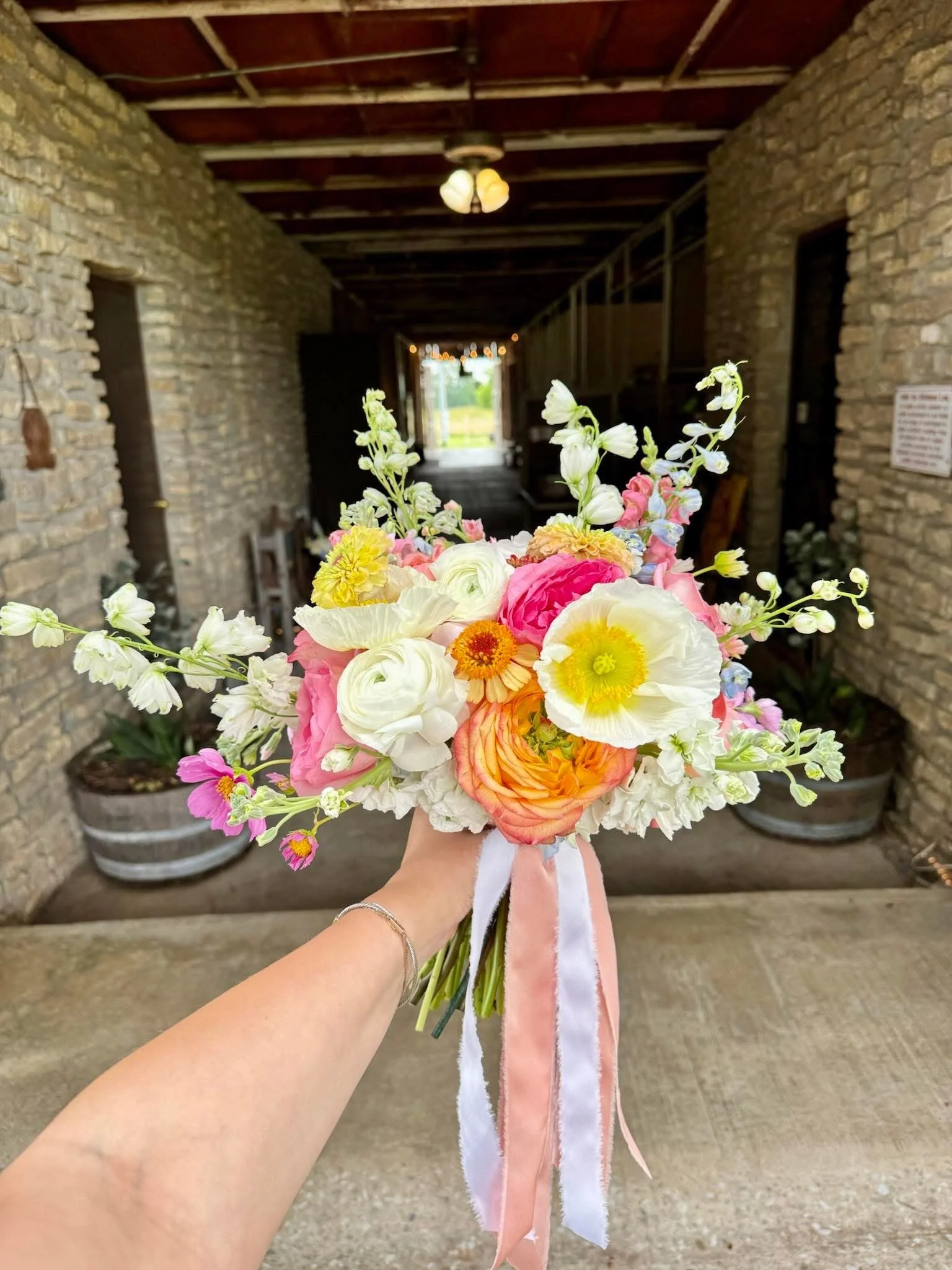 A person holding a colorful bouquet of flowers, with an arched brick wall and a doorway in the background.