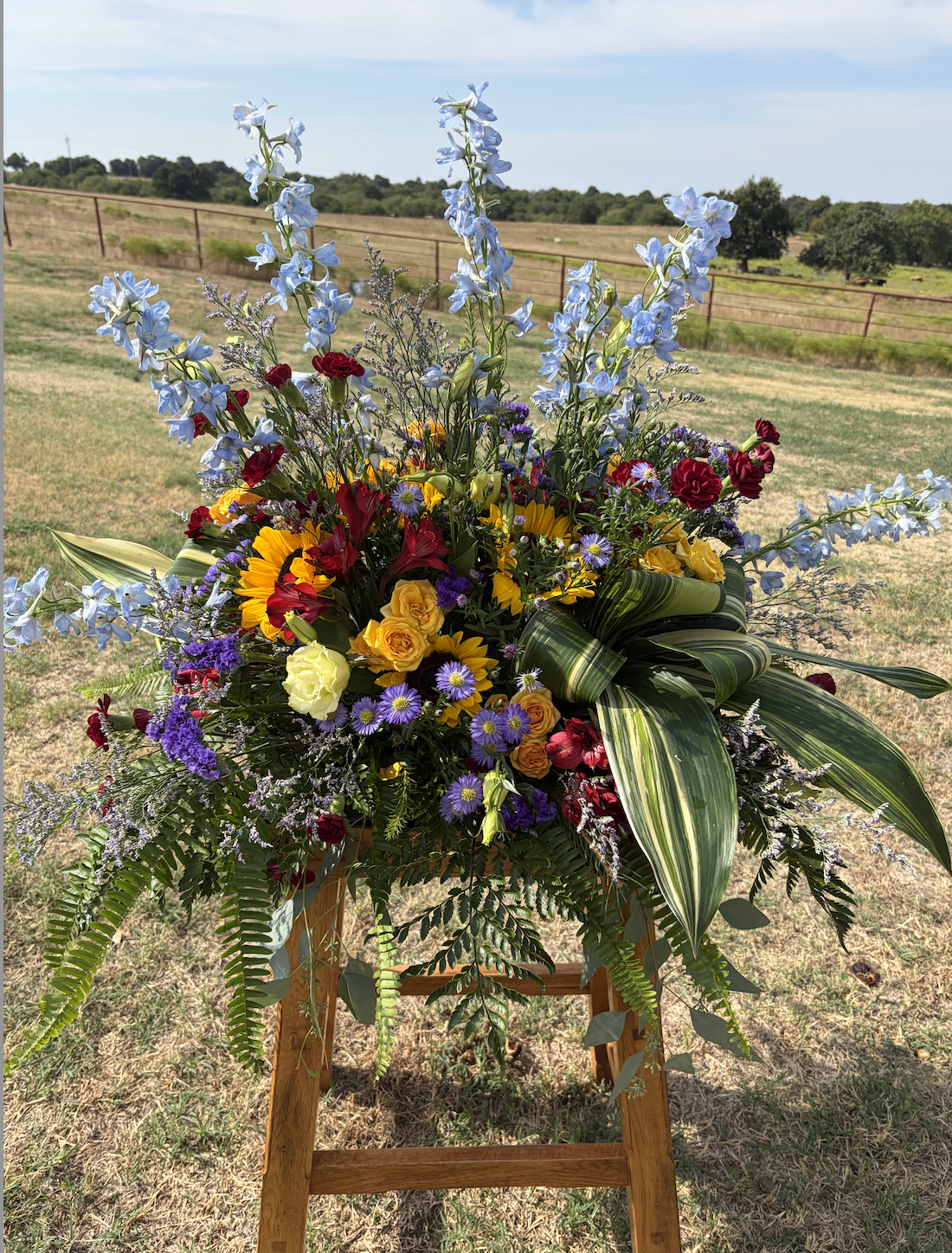 Colorful flower arrangement including yellow sunflowers, red roses, purple daisies, blue delphiniums, and green foliage, displayed outdoors on a wooden stand with a background of grassy field and trees.