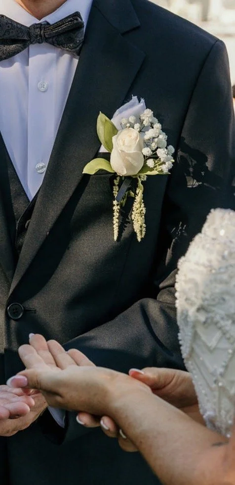 Close-up of a man in a black tuxedo with a white dress shirt and a bow tie, holding hands with a woman dressed in white, during a wedding ceremony. The man has a boutonnière with white roses and baby's breath on his suit lapel.