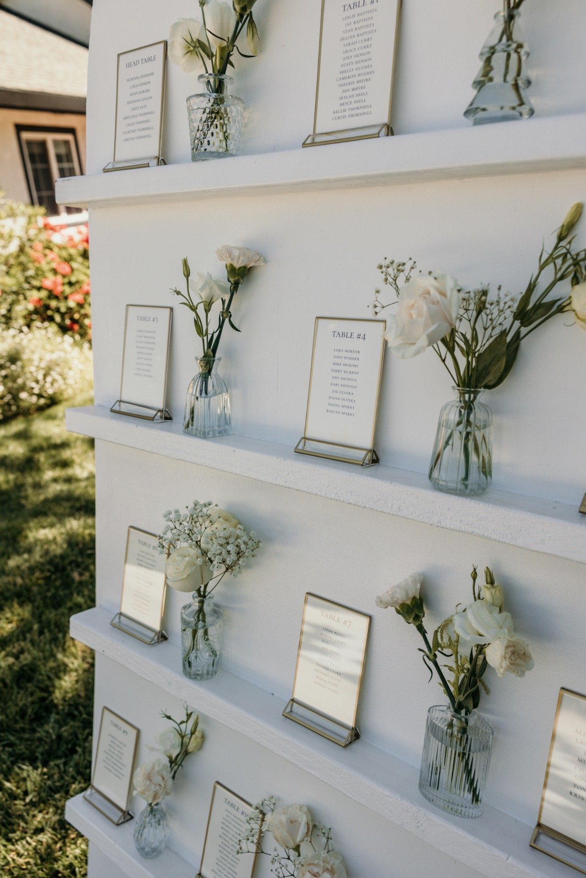 White wedding or event seating chart display with tables and guest names, decorated with white flowers in glass vases, outdoors with a grassy area and flowers in the background.