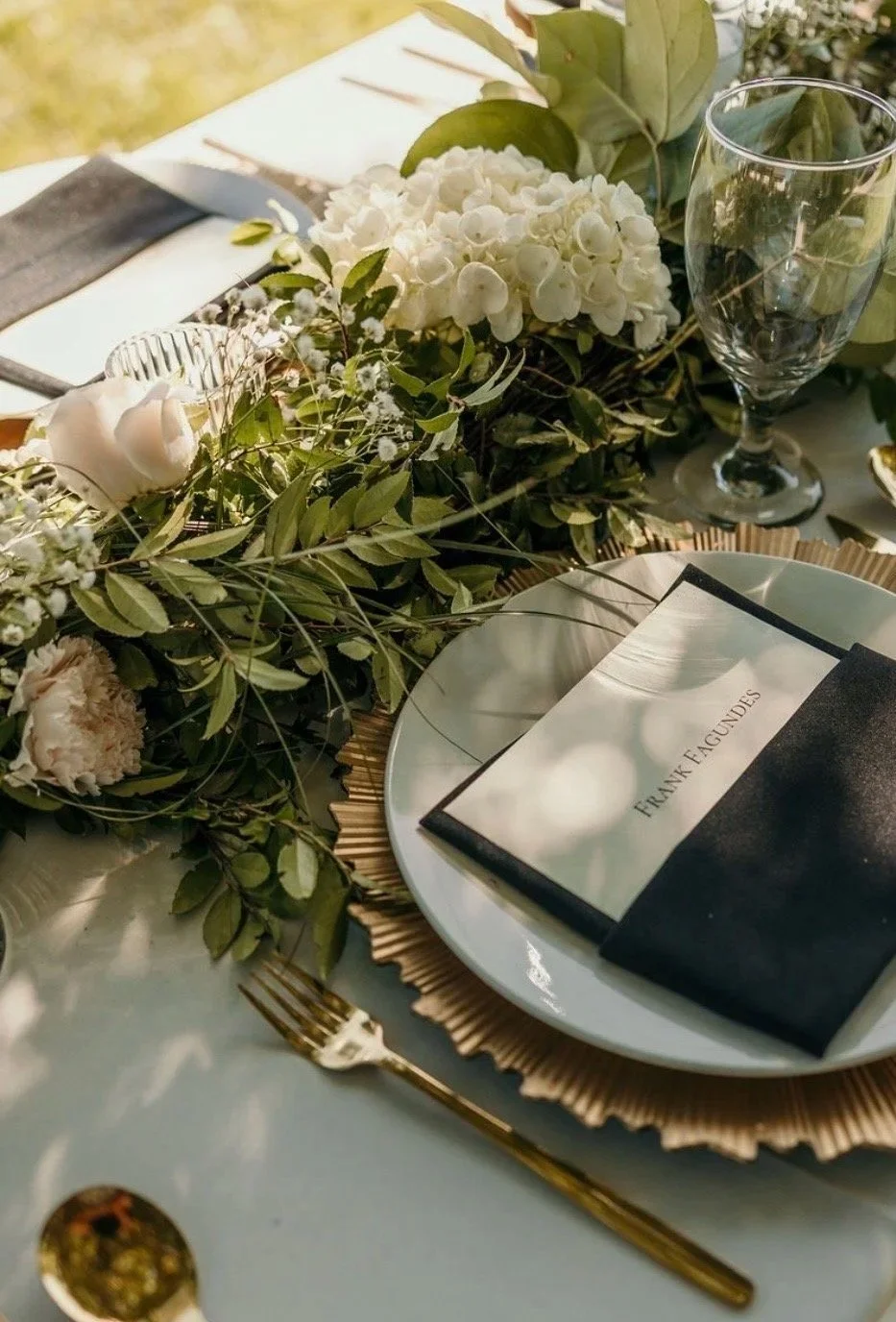 Elegant table setting with floral centerpiece, white hydrangeas, greenery, empty wine glass, patterned plate with black and white napkin labeled 'Frank Faucundes', and gold utensils.
