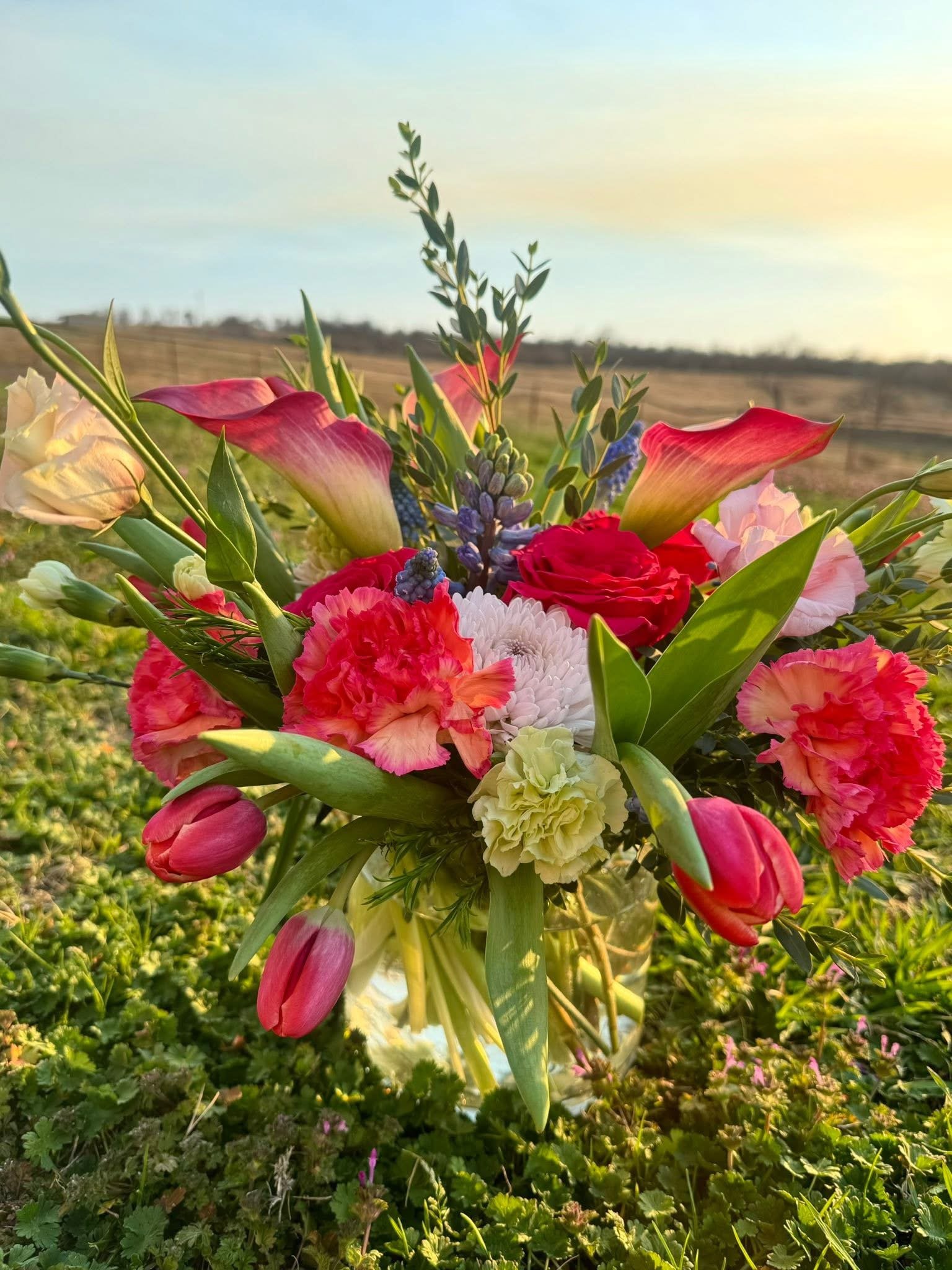 A bouquet of colorful flowers including calla lilies, roses, and other mixed flowers in a clear glass vase outside with a field and blue sky in the background.