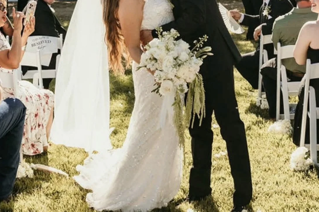 Wedding ceremony outdoors with a bride in a white dress holding a bouquet and a groom in a black suit. Guests are seated on white chairs on grass, and some guests are taking photos.