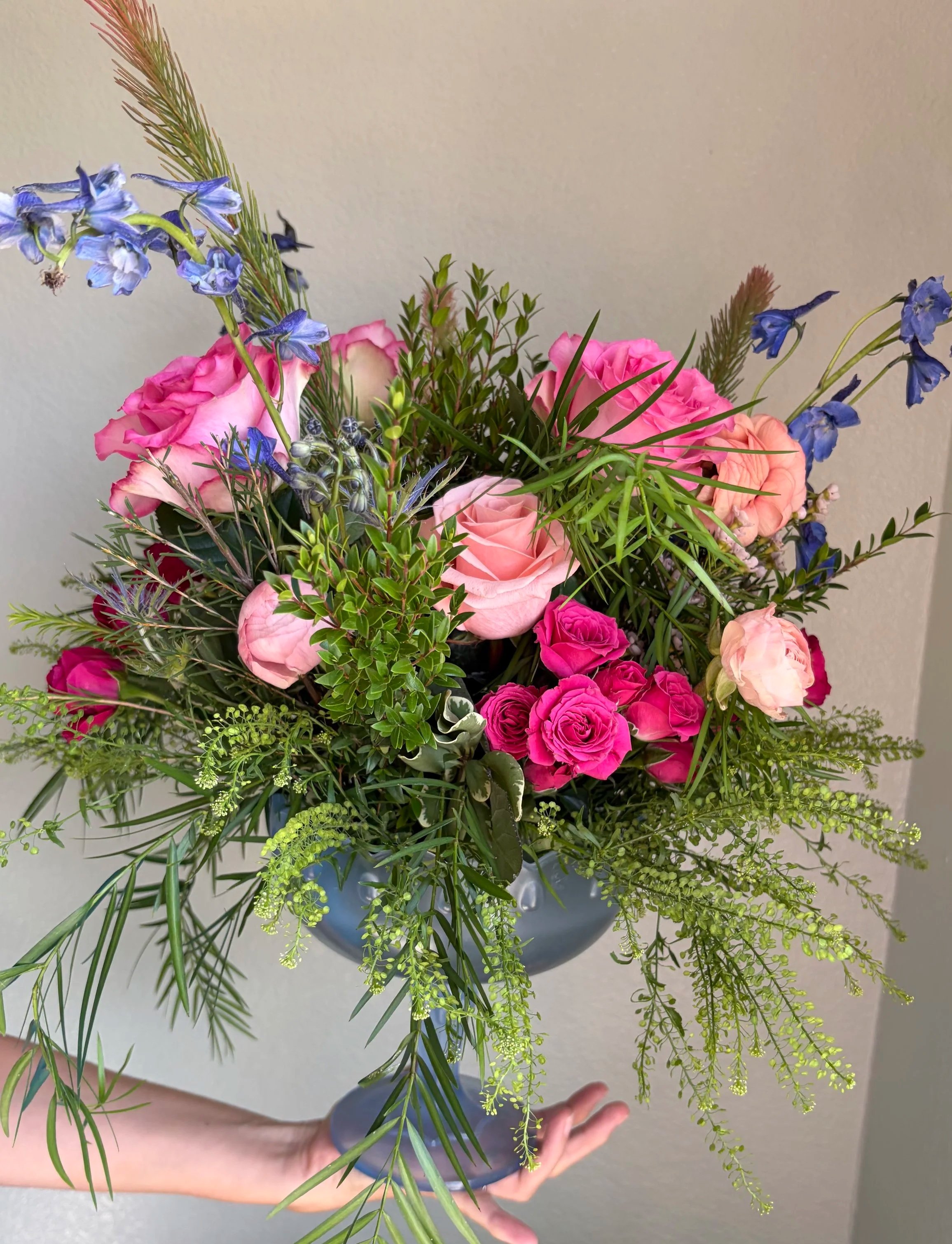 A person holding a deep blue vase filled with a colorful assortment of pink, purple, and yellow flowers, along with green foliage, against a plain gray background.