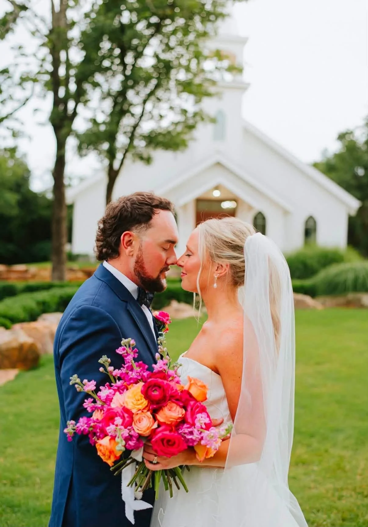 A bride and groom stand close together outdoors, with eyes closed, forehead touching, and noses almost touching in a romantic pose. The bride holds a large colorful bouquet of pink, orange, and yellow flowers. Behind them is a white church and green trees, suggesting a wedding setting.