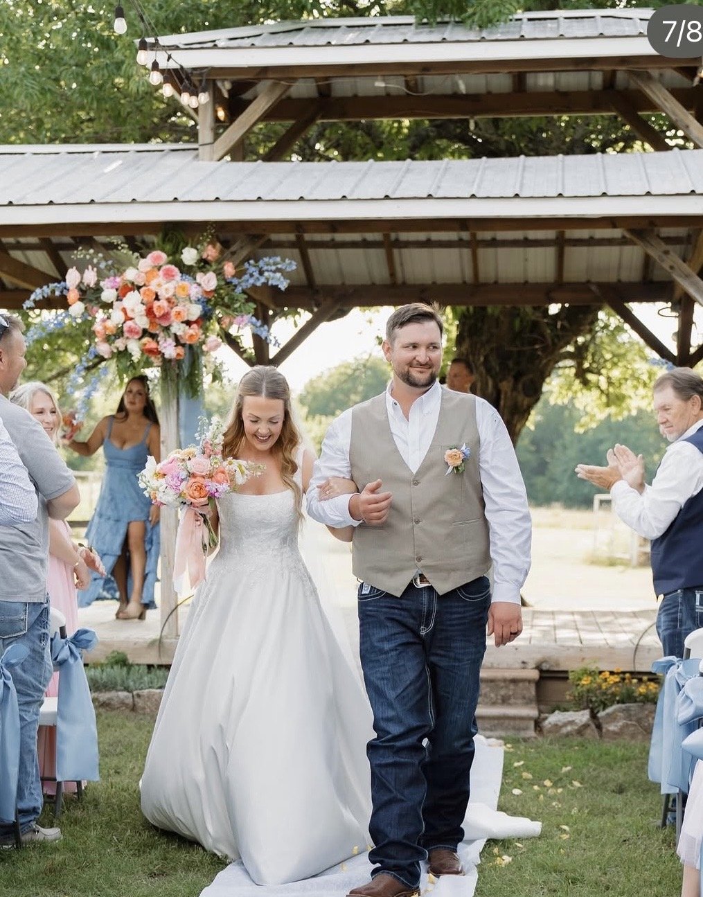 A bride and groom walking down the aisle at their outdoor wedding ceremony, with guests applauding and a floral arrangement in the background.