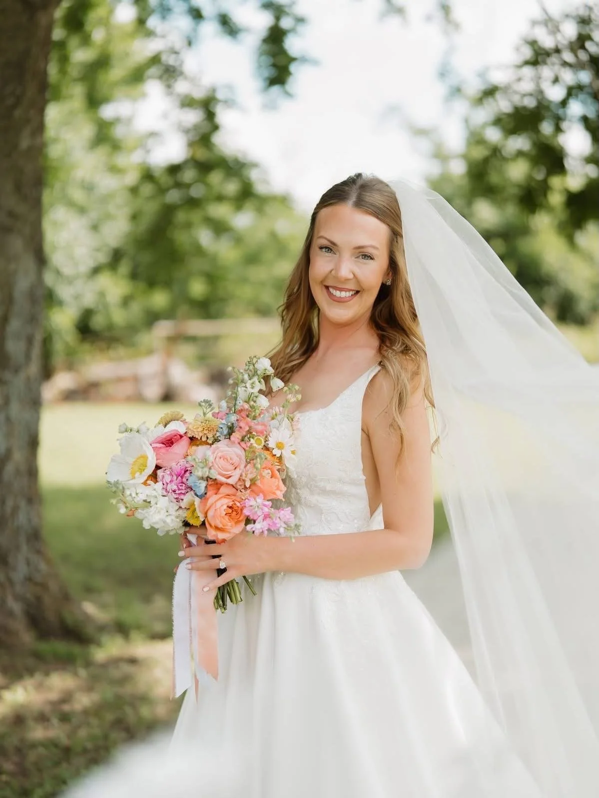 A smiling bride in a white wedding dress holding a colorful bouquet of flowers outdoors.