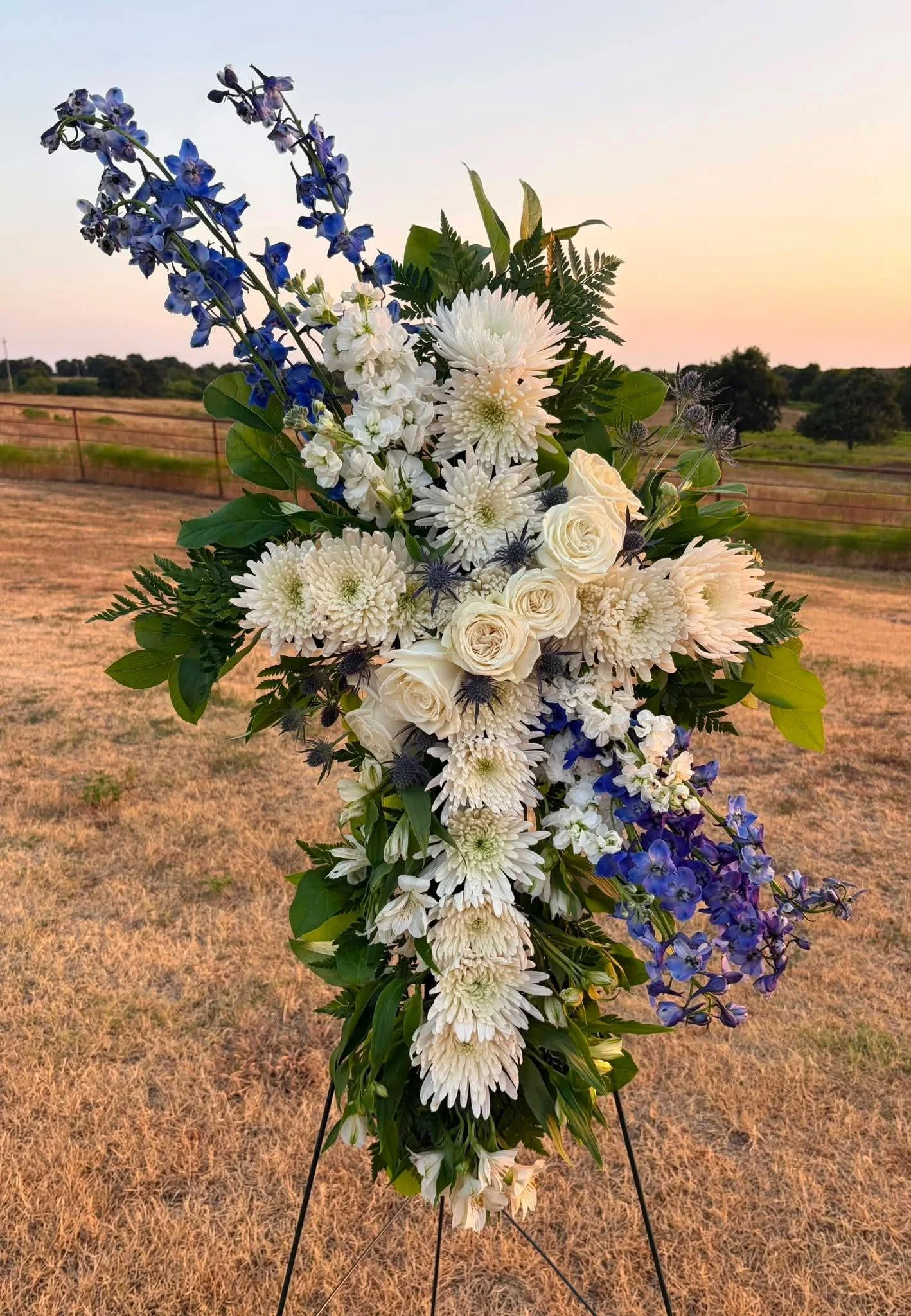 A bouquet of white, blue, and purple flowers with green foliage on a stand outdoors during sunset with a field and trees in the background.