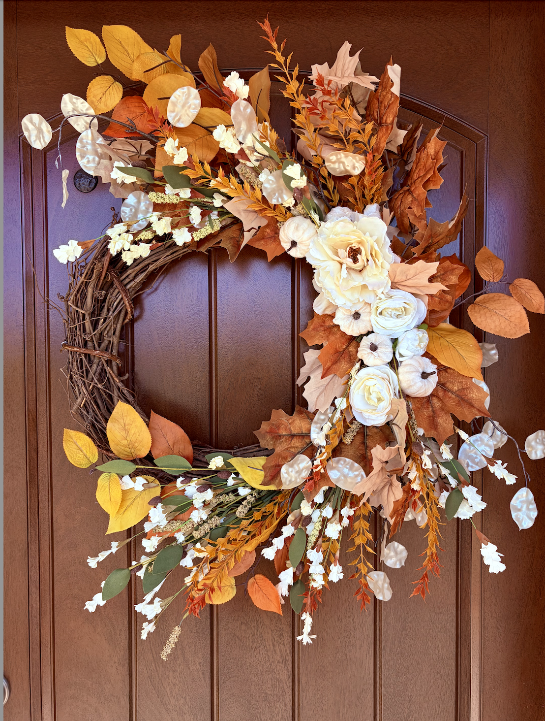 Autumn-themed door wreath with white flowers, orange and brown leaves, and decorative pearl-like accents, hanging on a wooden door.
