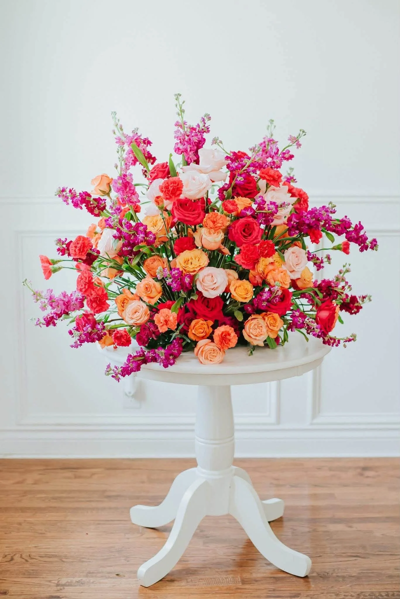 A colorful bouquet of mixed flowers, including roses and carnations, on a white pedestal table in a room with light-colored walls and wooden floor.