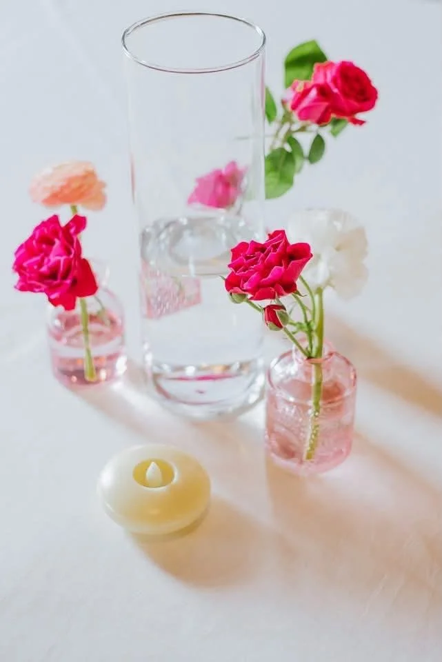 Arrangement of pink, white, and orange flowers in small vases, with a clear glass vase filled with water and a candle on a light-colored surface.