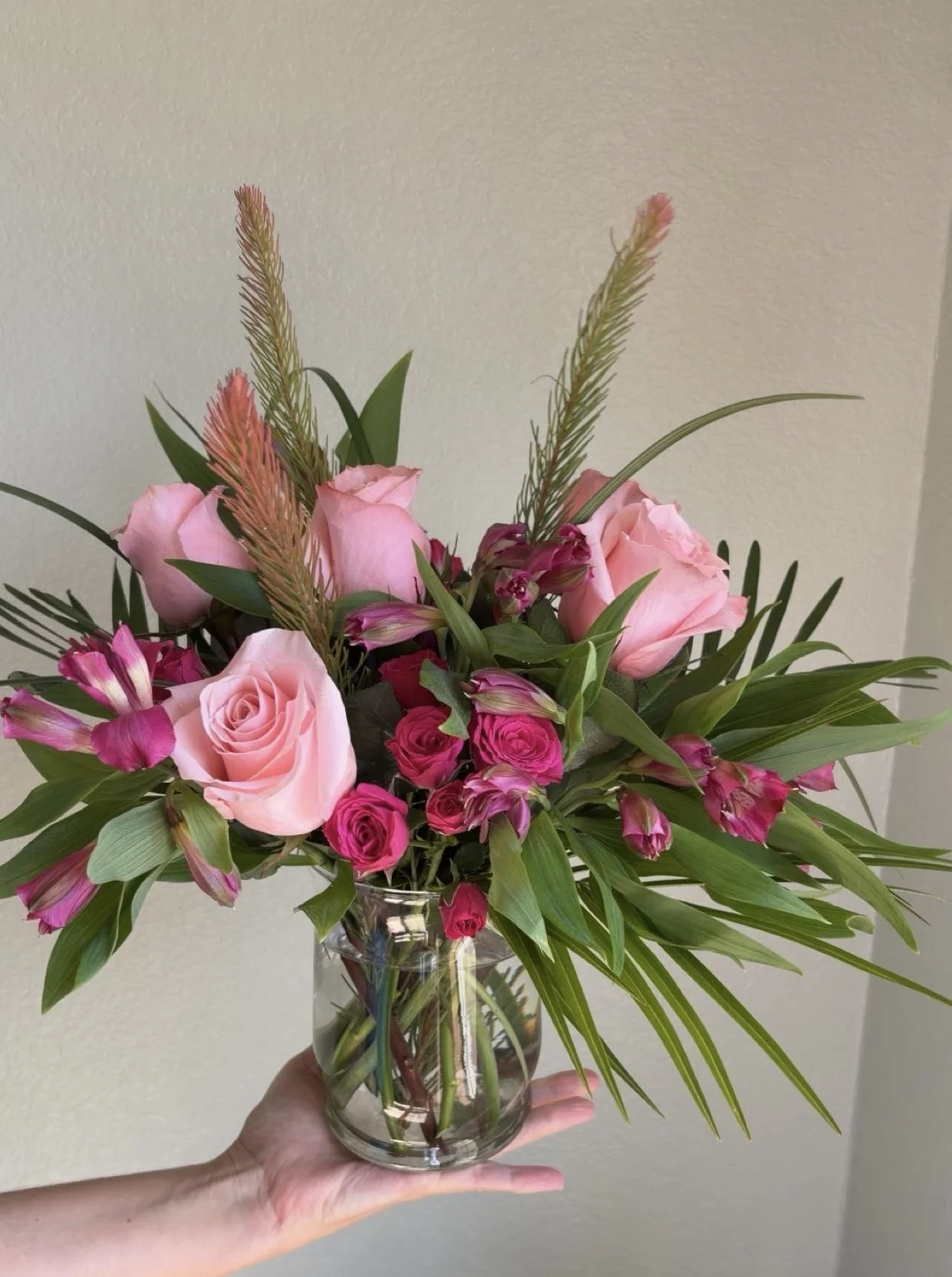 A hand holding a clear glass vase with a pink and purple flower bouquet, including roses, lilies, and tall grasses, against a plain wall.