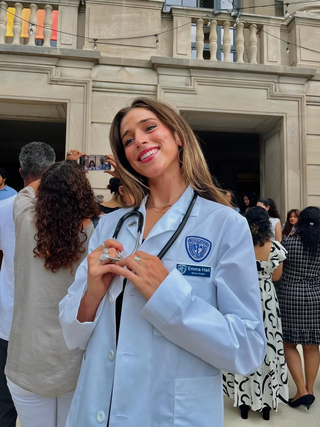 A young woman in a white medical coat with a stethoscope around her neck, smiling and holding her coat at the chest, surrounded by people at what appears to be a graduation or ceremony event.