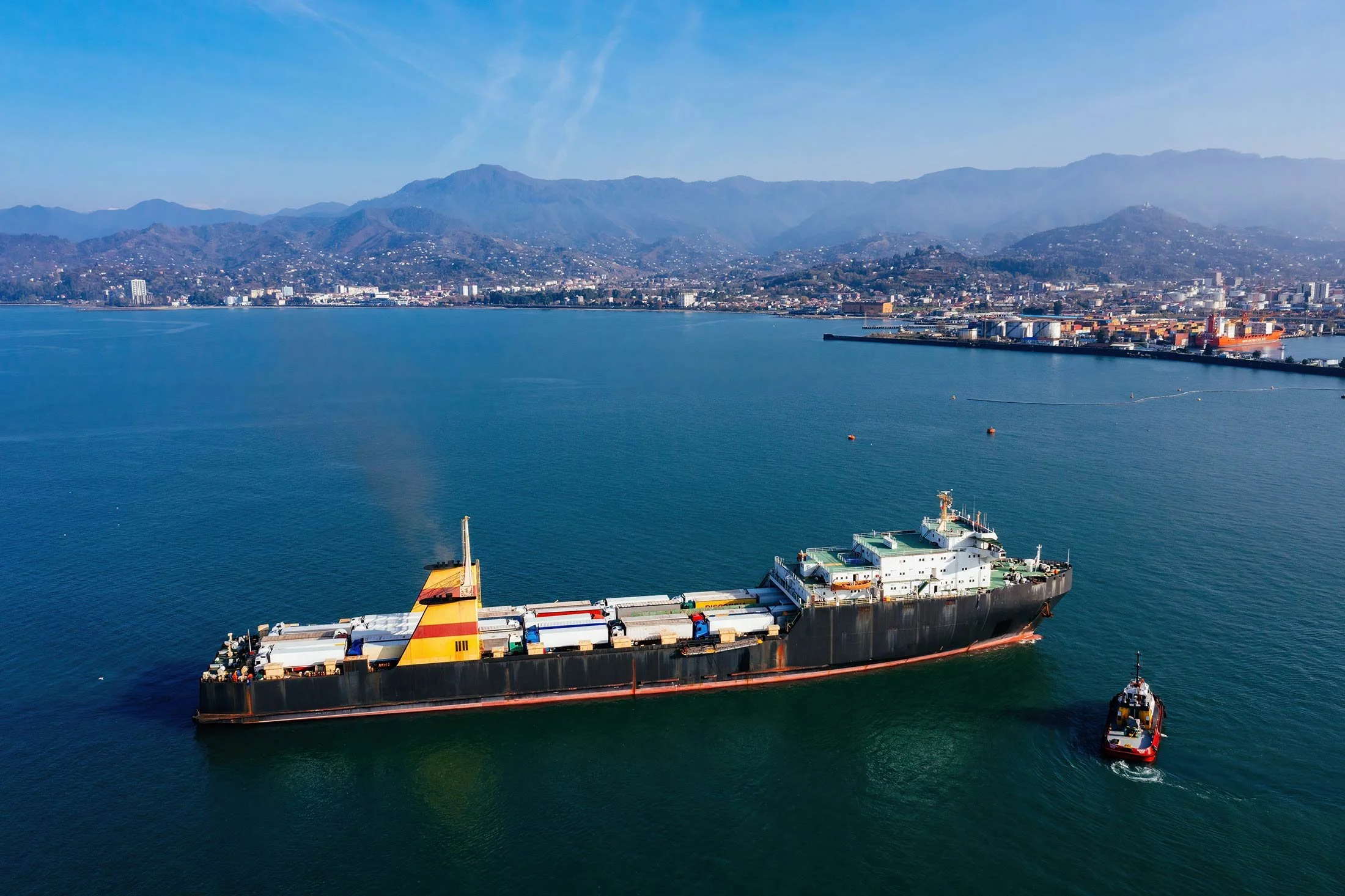 A cargo ship with a tugboat in a harbor, with a city and mountains in the background on a sunny day.