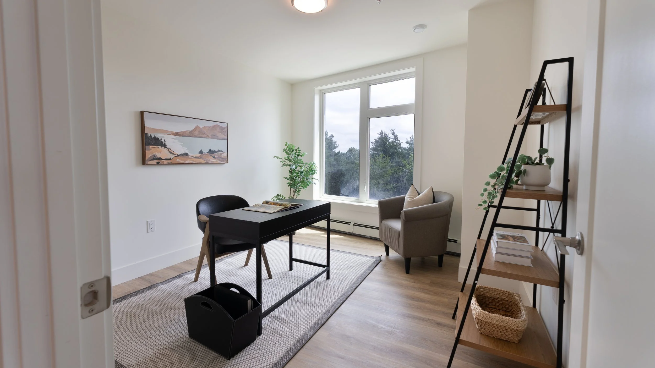 Professional Halifax Nova Scotia real estate photography. Room with large window showing trees outside, black desk with open book, grey armchair, black chair, potted plant, bookshelf with books and decor, and a framed landscape painting on the wall.