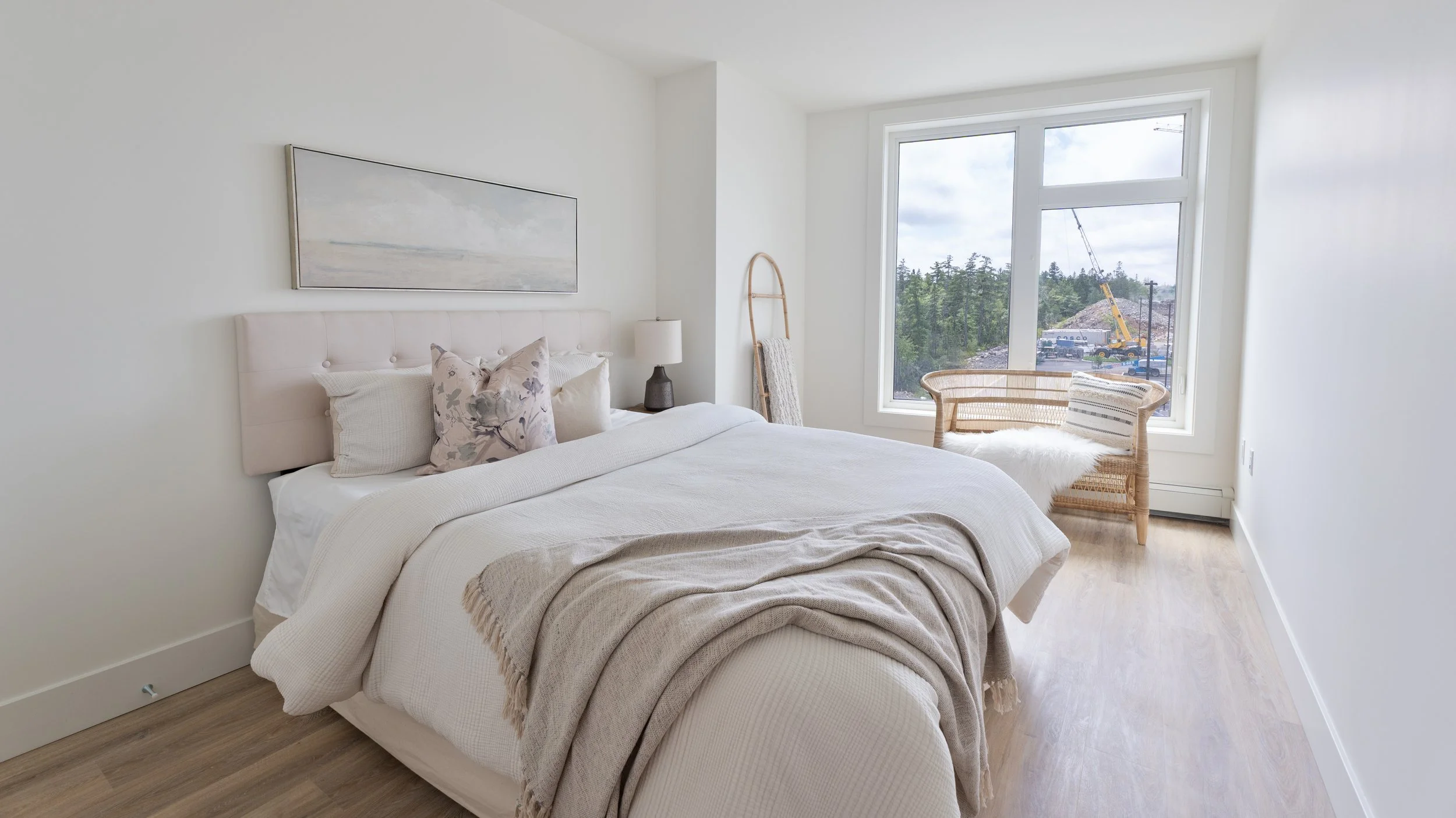 High quality Halifax real estate photography. MLS listing photography. Bedroom with a white bed, beige blanket, decorative pillows, a nightstand with a lamp, a wicker chair with a fur throw, and a large window.