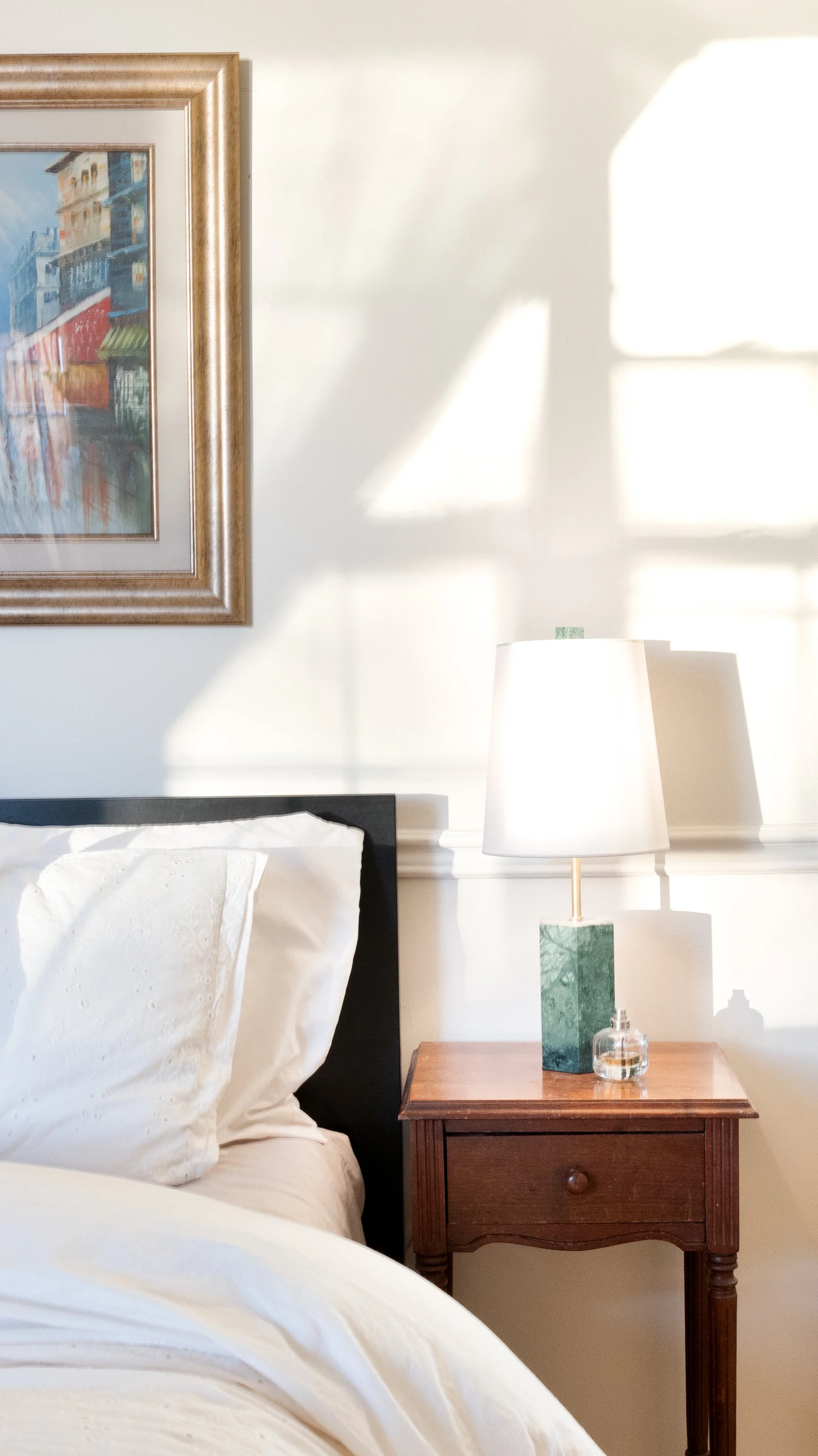 Interior Photography. A close-up of a bedroom with a bed, white bedding, a wooden nightstand, a white lampshade with a green marble base, a small glass perfume bottle, and a framed painting on a white wall with sunlight casting geometric shadows.
