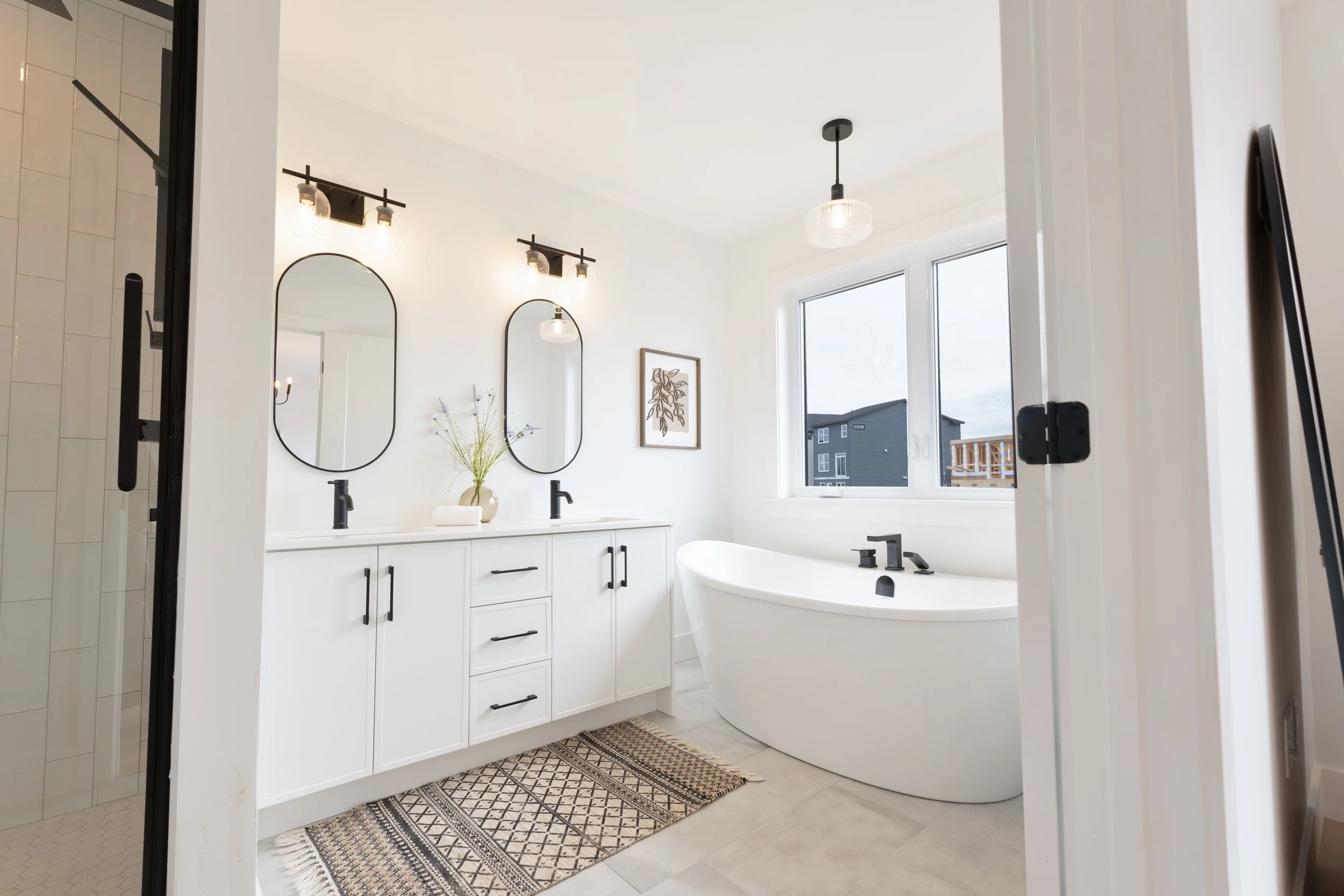 High quality Halifax real estate photography. MLS Listing Photography. Modern white bathroom with double vanity, black fixtures, oval mirrors, a freestanding bathtub near the window, a patterned rug, and a shower stall with glass door on the left.