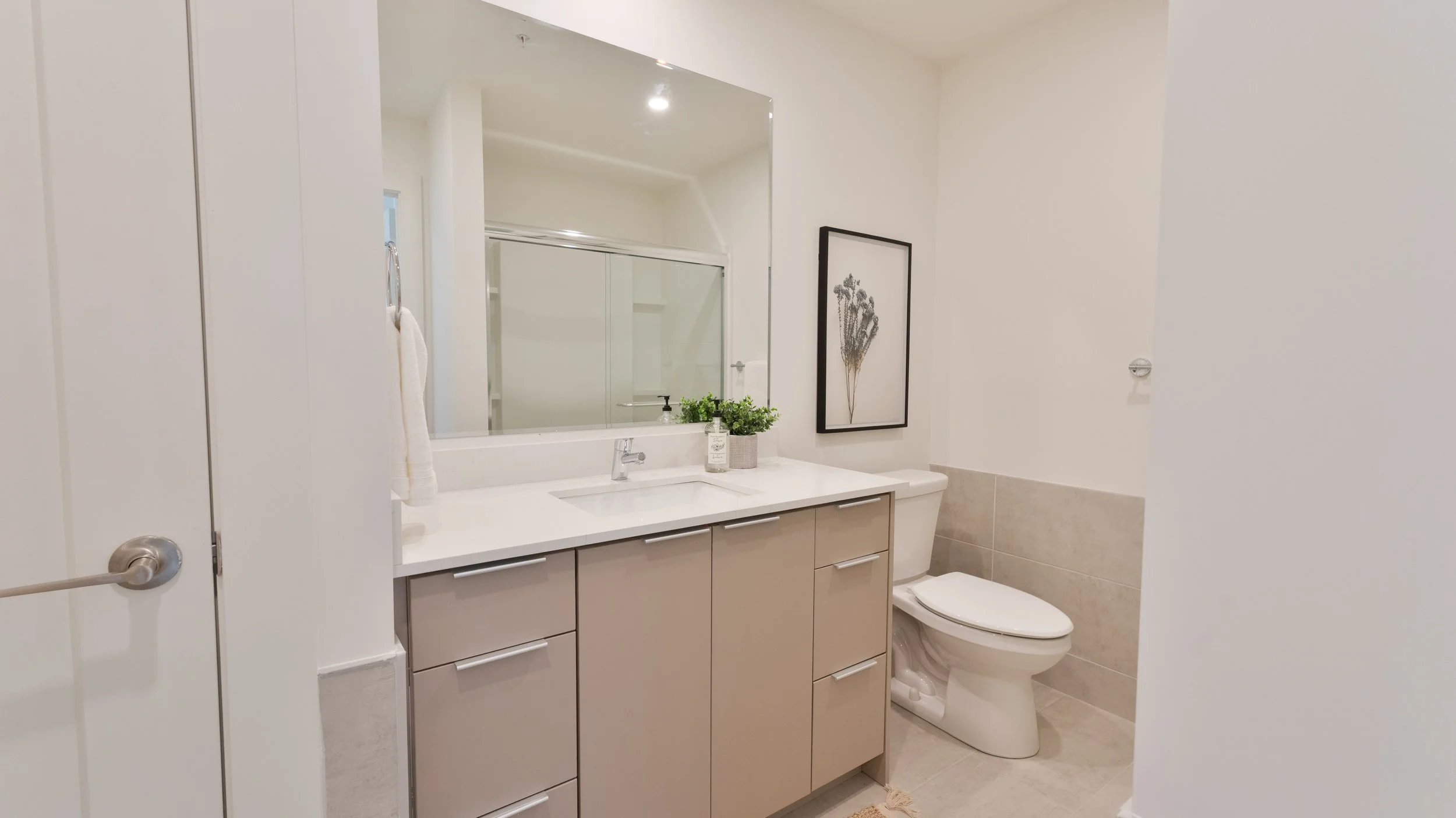 Professional high quality Halifax Nova Scotia real estate photography. MLS Listing Photo. Modern bathroom with beige vanity, white countertop, large mirror, towel ring with towel, decorative plants, framed art, toilet, and neutral-colored tiles.