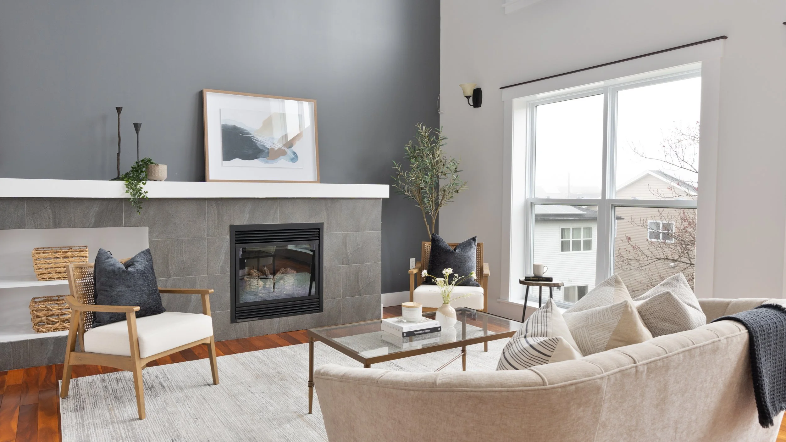 Living room with a light-colored sofa, a glass coffee table, a cozy armchair with a black pillow, and a large window letting in natural light. There is a fireplace with a dark gray wall behind it, decorated with a framed artwork, plants, and candles.
