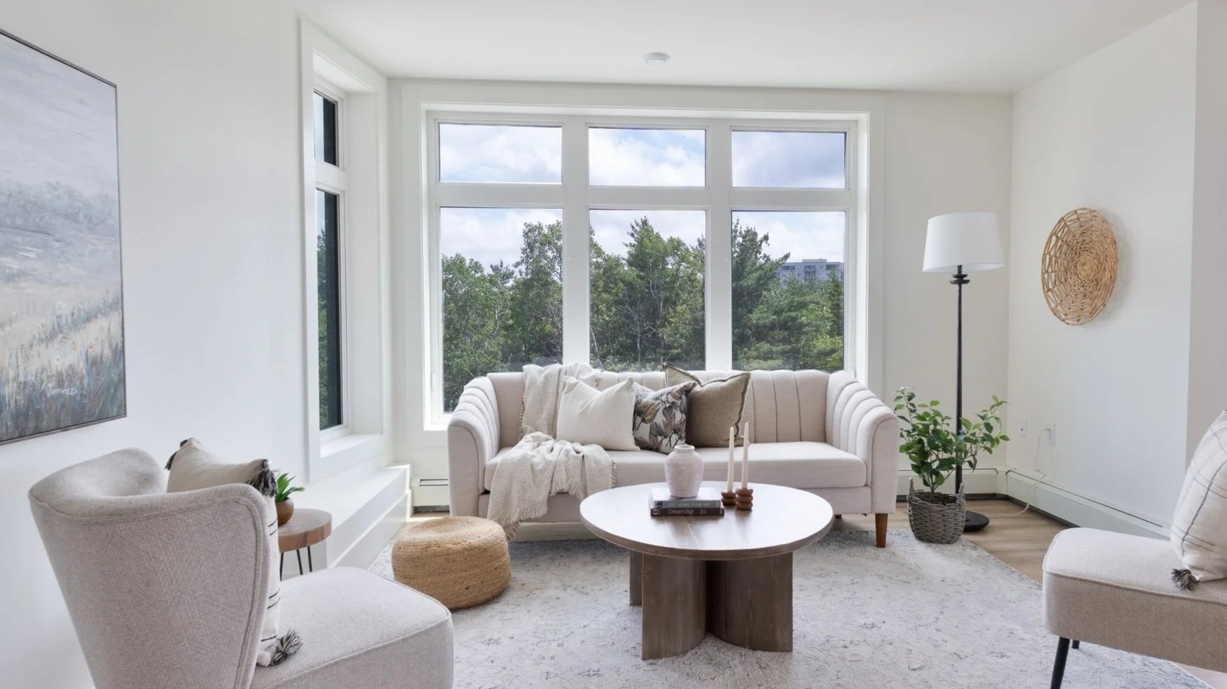 Living room with white walls, large windows, and neutral furniture including a beige sofa, white armchair, and round wooden coffee table decorated with candles, books, and vases. There is a floor lamp, a plant in a woven basket, and wall art.