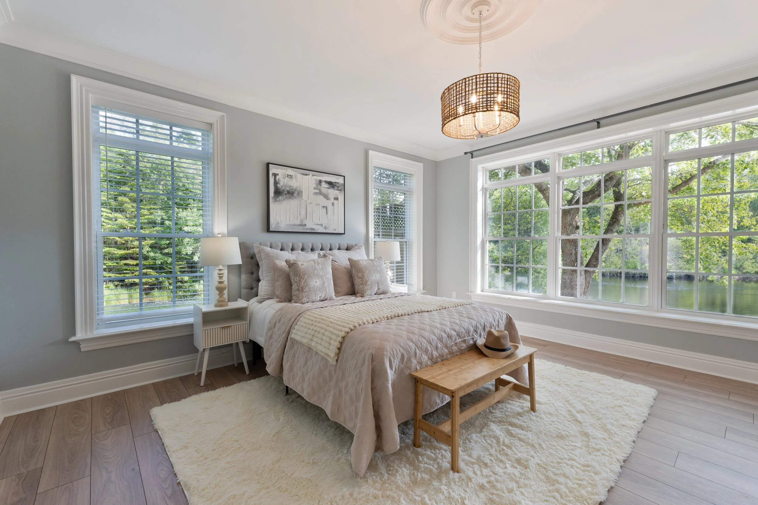Nova Scotia, Halifax real estate photography and marketing for MLS listing.A bedroom with large windows overlooking trees and water, a bed with beige bedding and pillows, two bedside lamps, a wooden bench with a hat on it, and a large white shag rug.