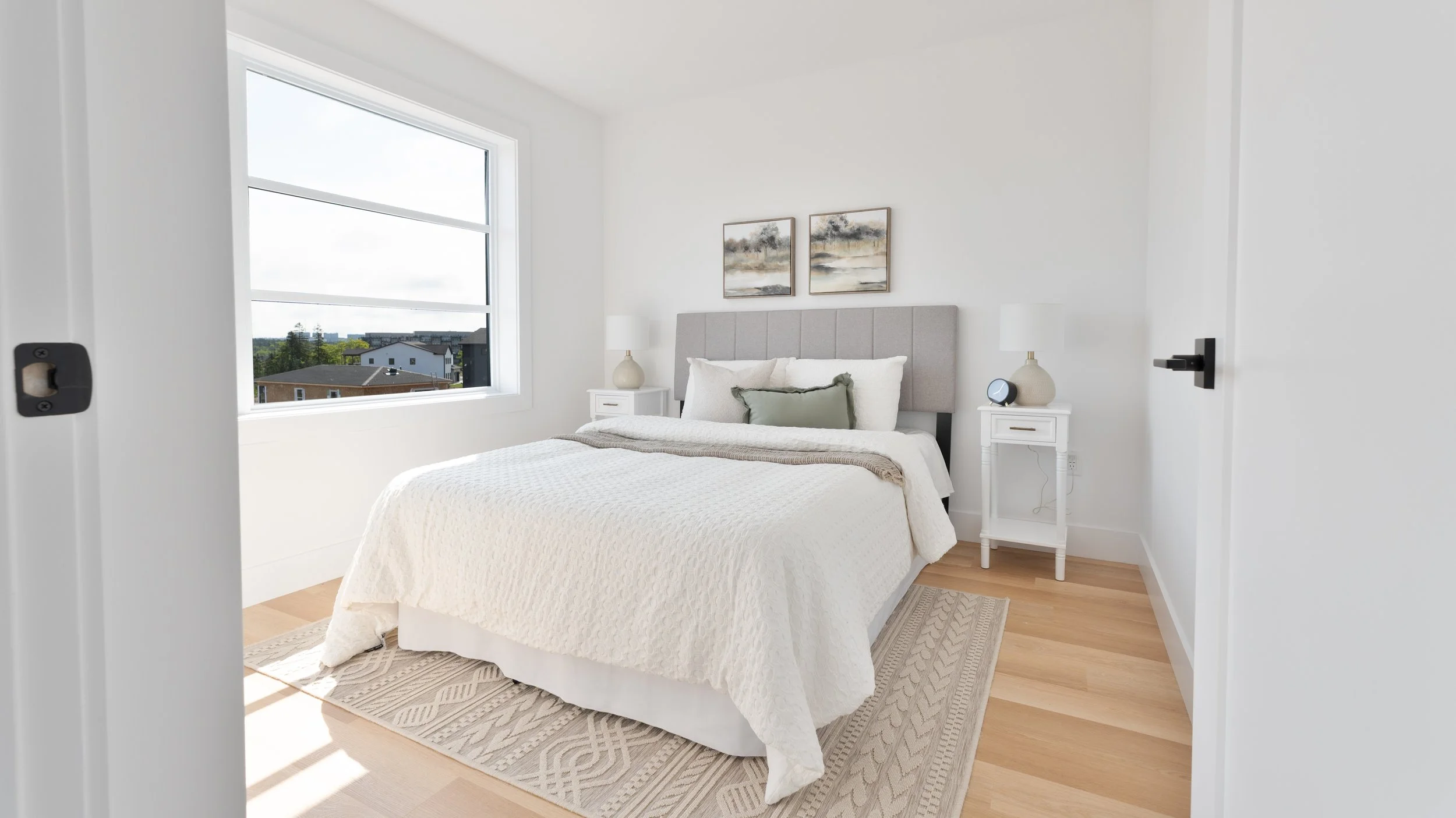 High quality Halifax real estate photography. A minimalist bedroom with white walls, a large window showing a suburban neighborhood, a bed with white bedding and decorative pillows, two white nightstands with lamps, and small artwork above the bed.