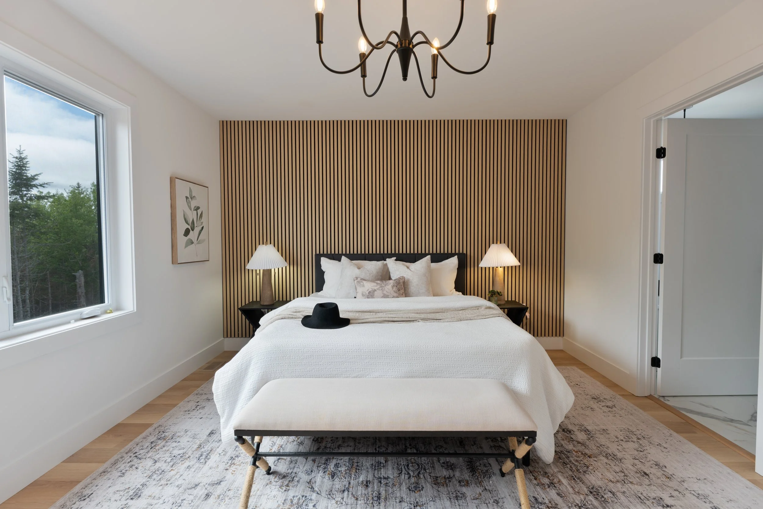 Nova Scotia, Halifax real estate photography for MLS. Shows modern bedroom with a white bed, black headboard, two bedside lamps, and a wooden slat accent wall. A black hat rests on the foot of the bed, and a bench is placed at the foot of the bed. 