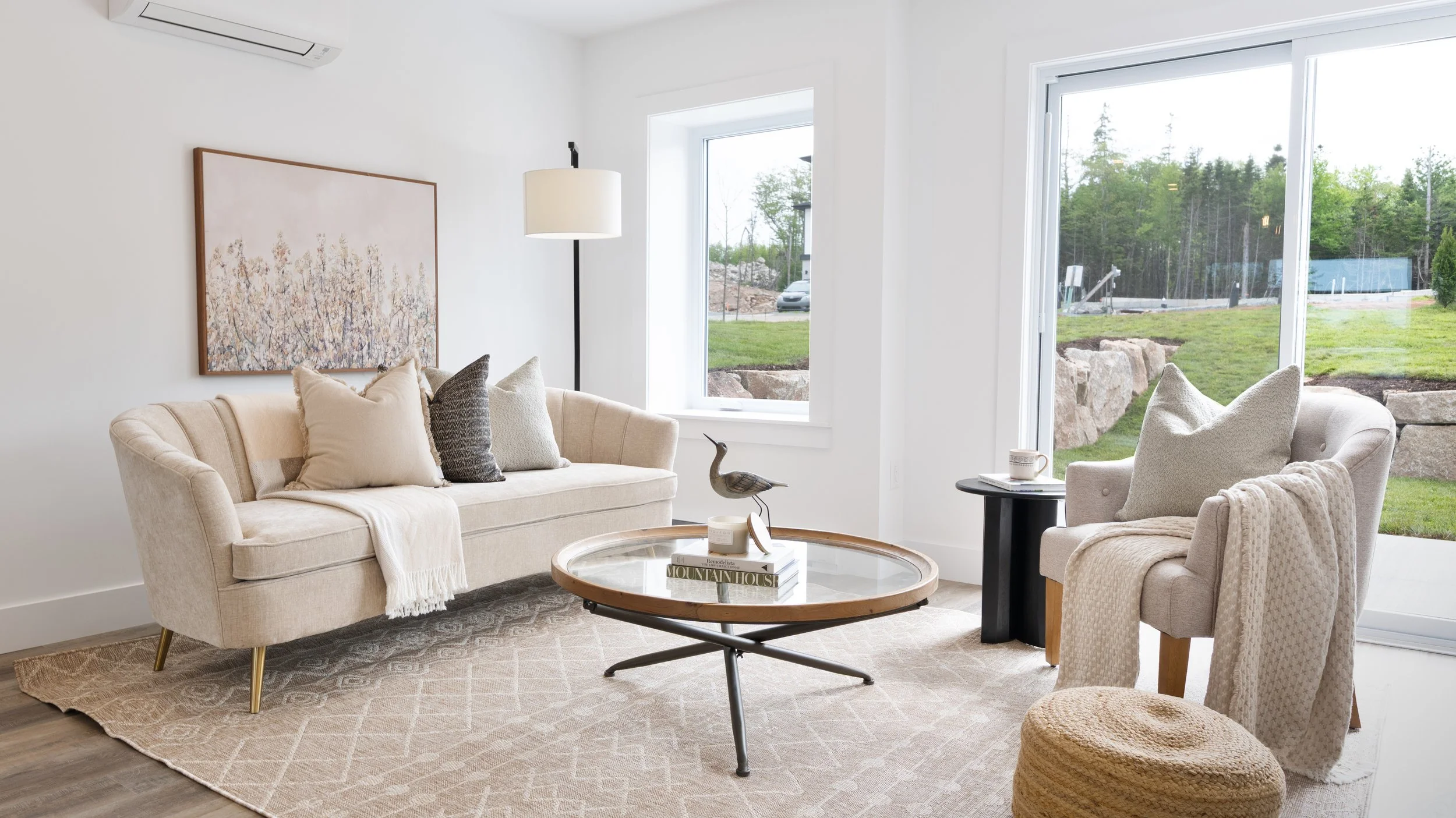 Professional high quality Halifax real estate photography. MLS Listing Photo A living room with a beige sofa, multiple throw pillows, a blanket, and a round glass coffee table with decorative items, large windows and a view of outdoor greenery.