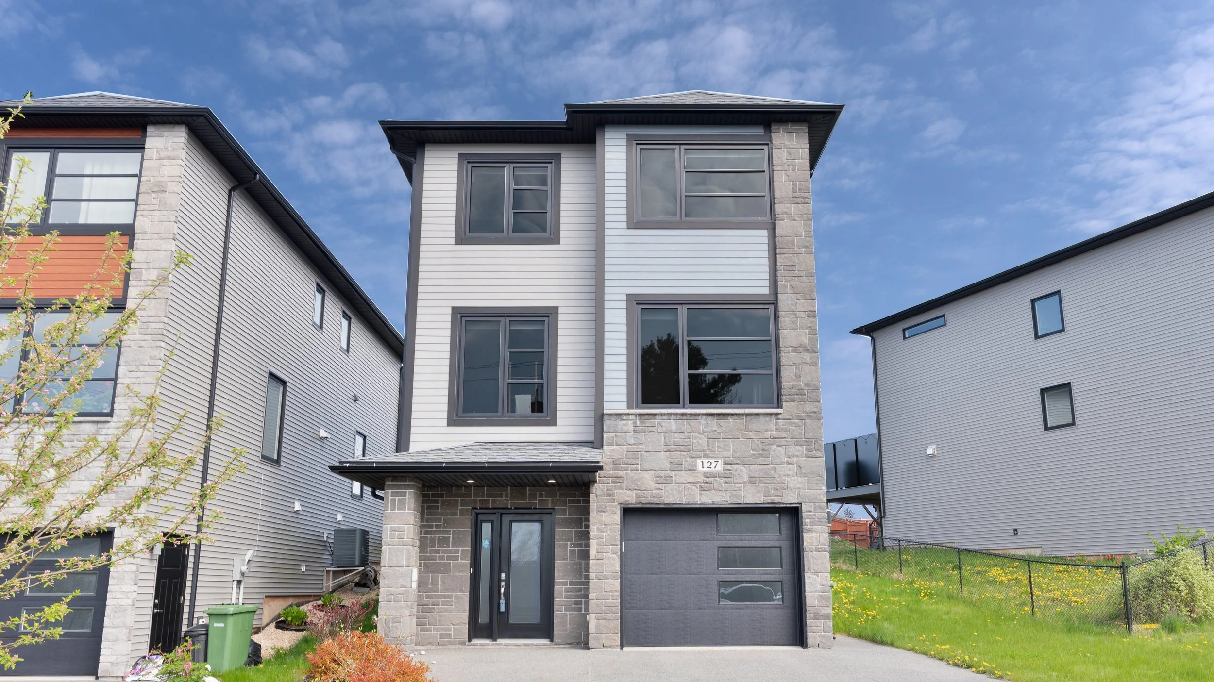 Nova Scotia, Halifax real estate photography for MLS. Exterior shot of standalone house with grey and white siding, large windows, and a stone facade at the entrance. There is a garage on the right side and a small front yard with grass and plants. 