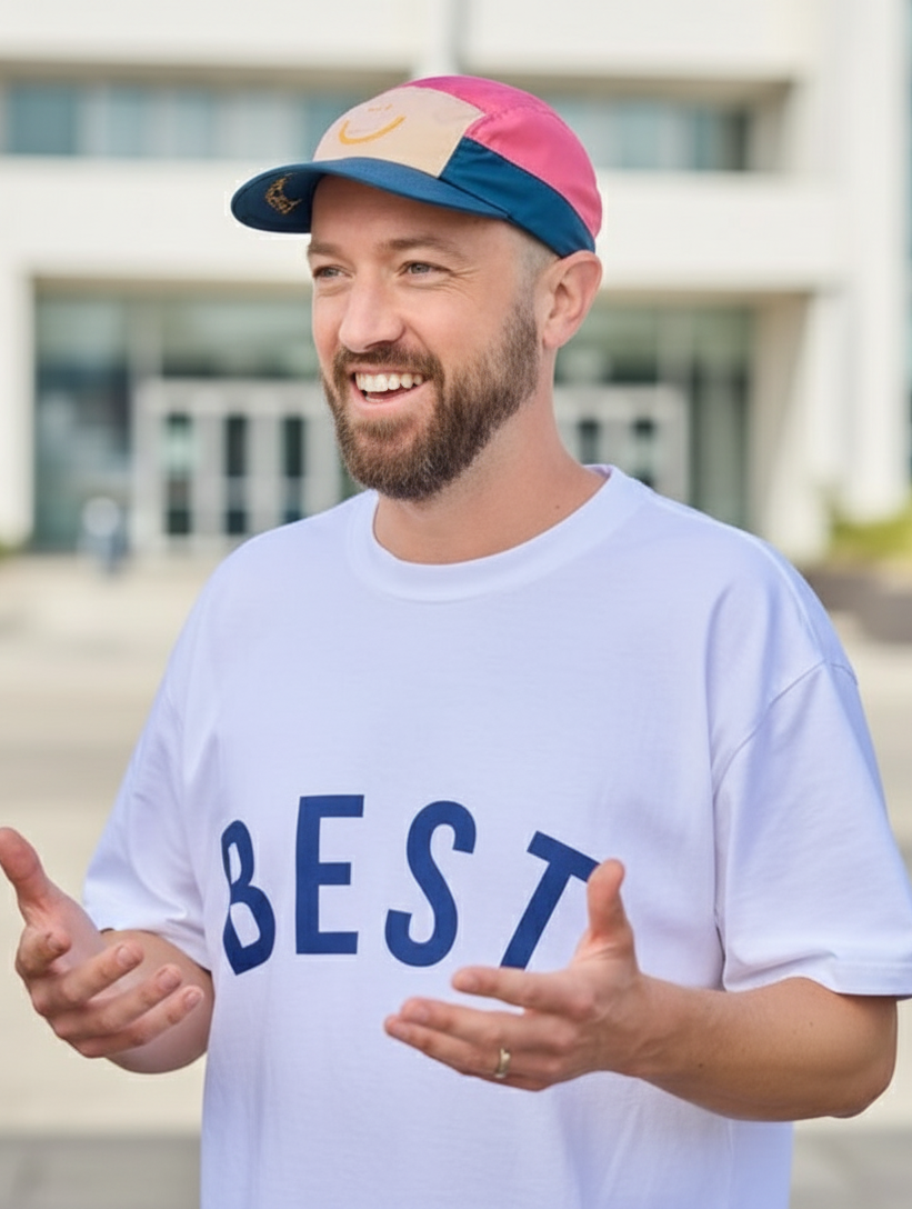 A man with a beard smiling outdoors, wearing a white T-shirt with the word "BEST" in blue letters and a pink, white, and blue cap.