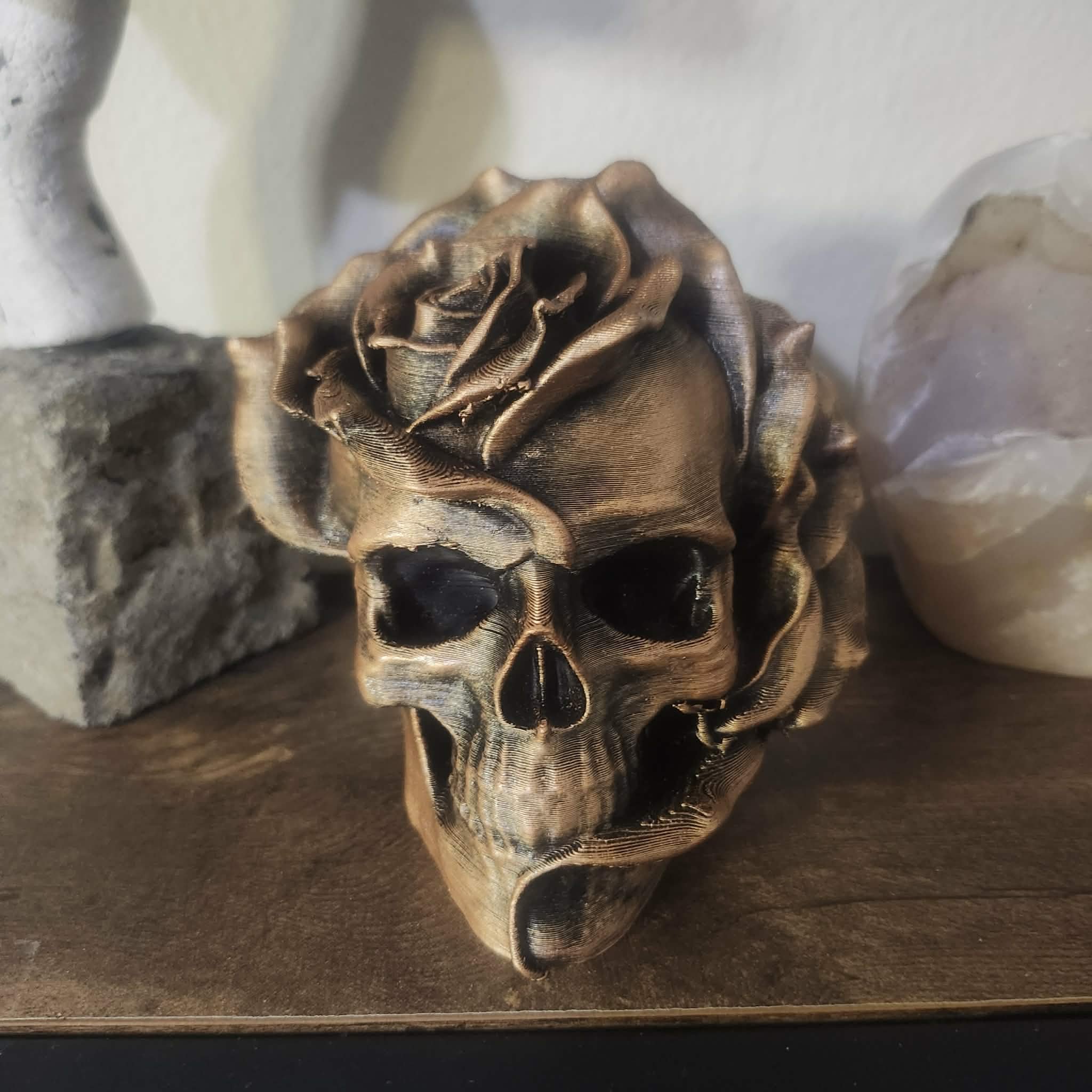 A decorative skull sculpture with a metallic finish, topped with a rose-shaped ornament, placed on a wooden surface.