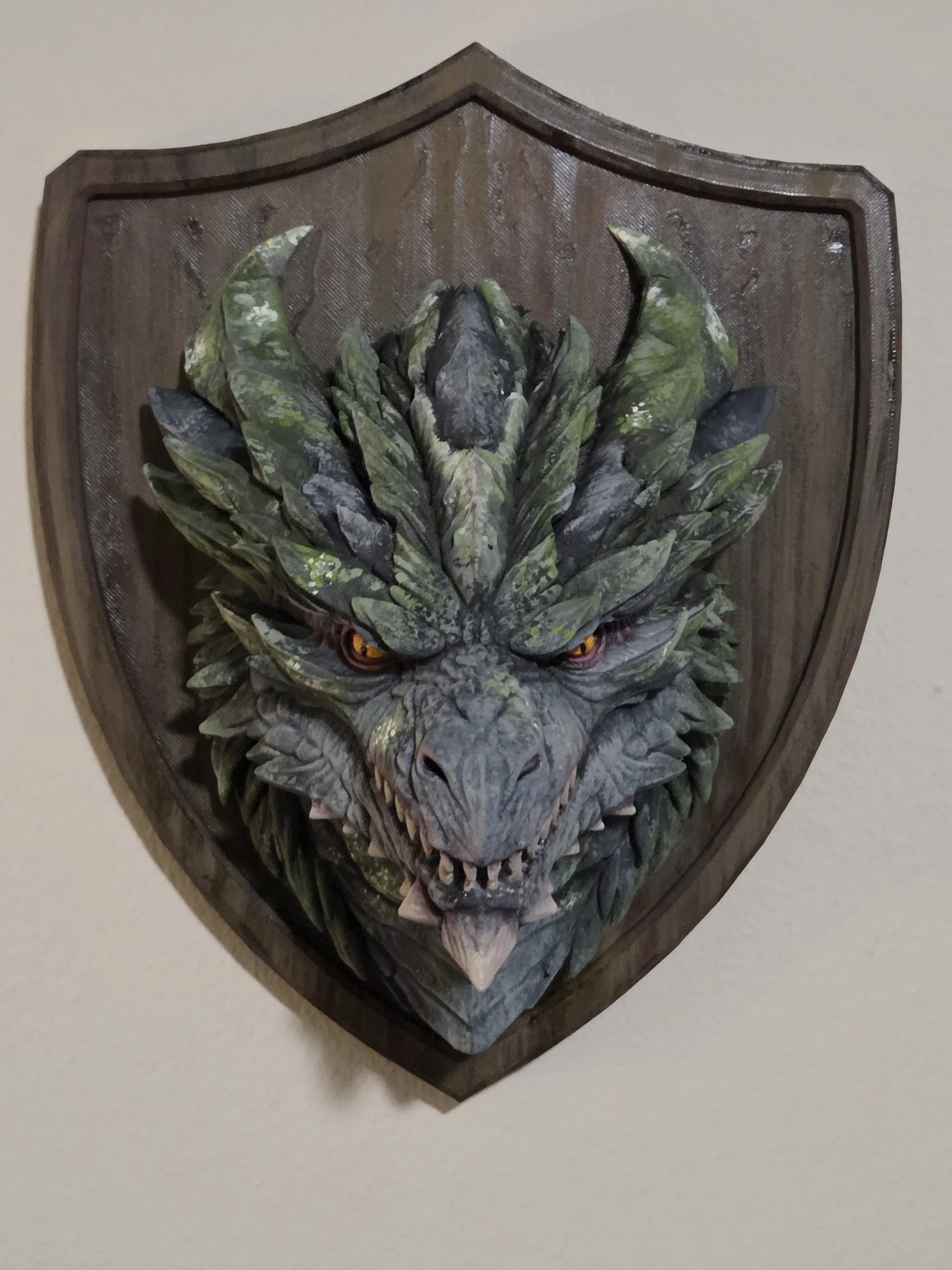 A detailed wooden plaque of a fierce dragon's head with green and gray scales, sharp teeth, and glowing orange eyes, mounted on a wall.