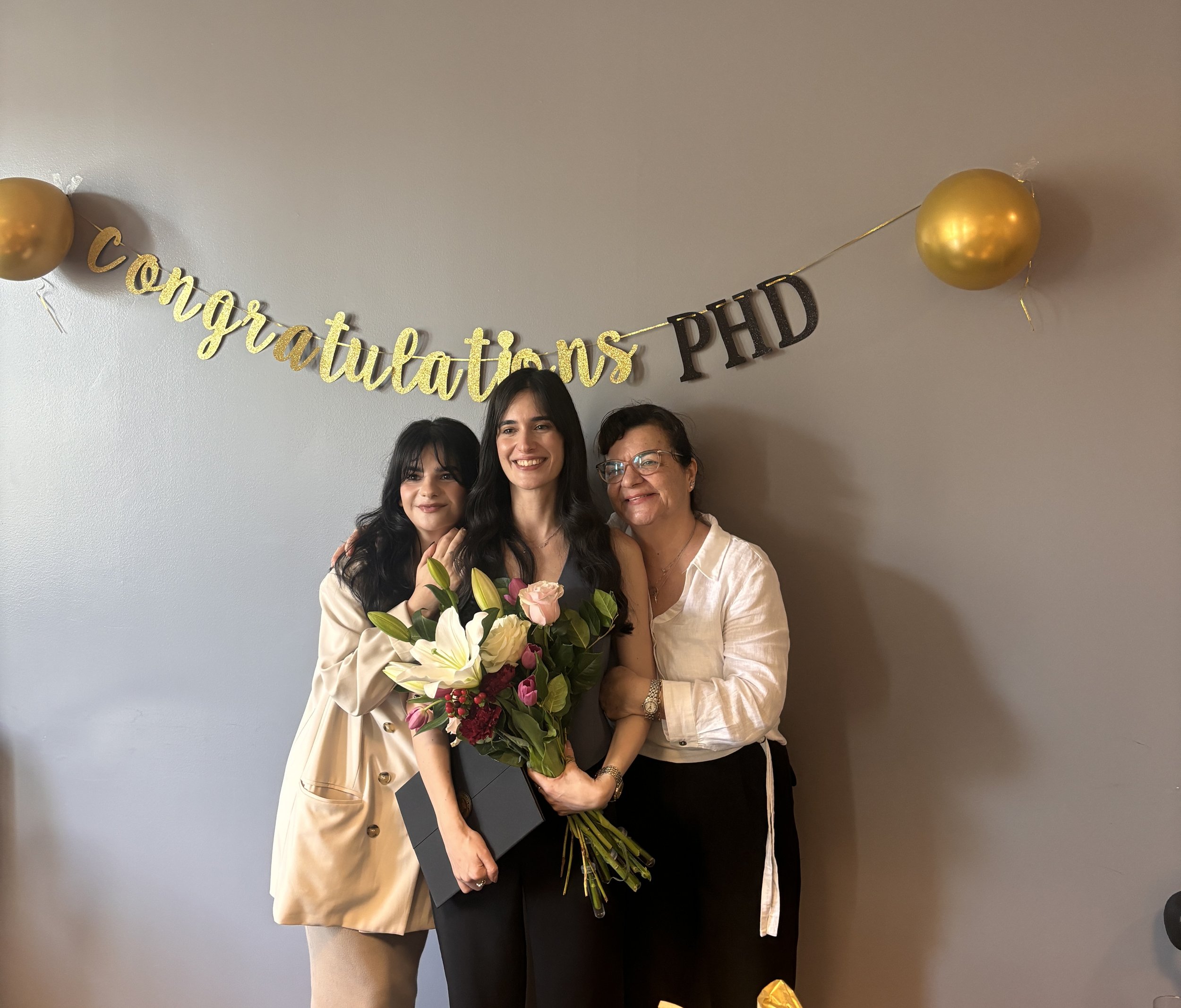 Three women celebrating graduation. One woman in the center receives a bouquet of flowers, smiling, with a gold and black banner reading 'Congratulations PhD' and gold balloons in the background.
